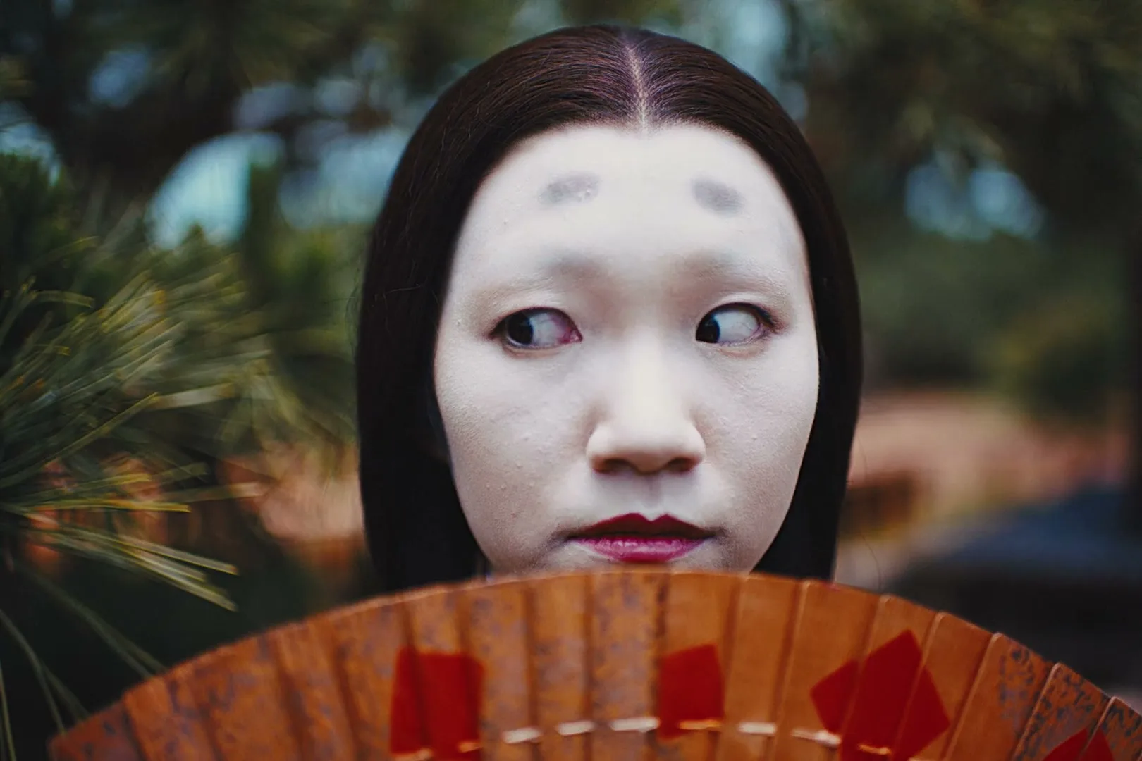 A young woman with traditional Japanese white face makeup and painted eyebrows looks around nervously, holding a fan in front of her mouth, outdoors.