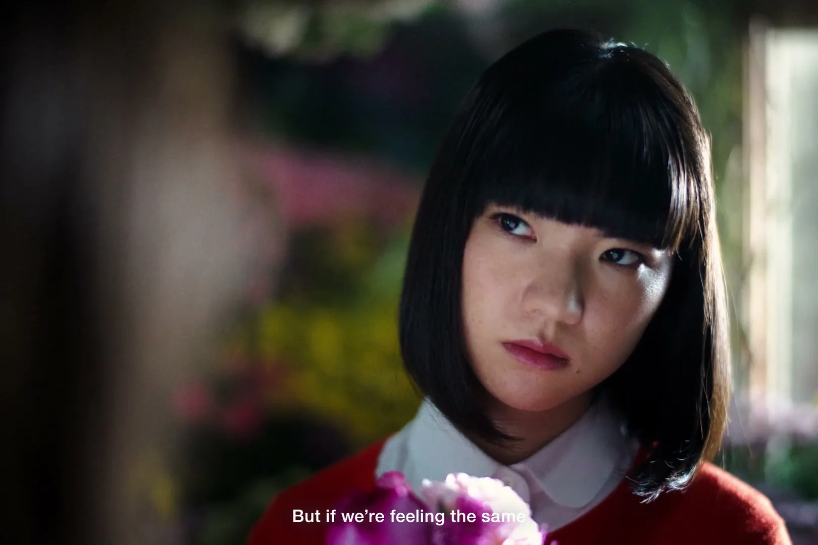A young Japanese woman with a bob haircut and a red sweater looks up with a concerned expression, holding a pink flower indoors.