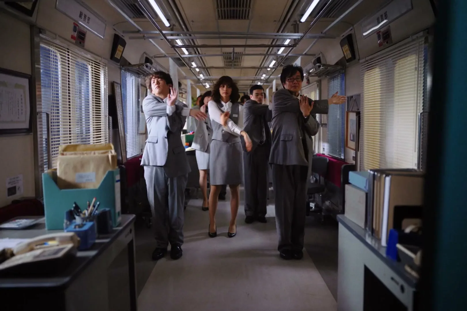 A young woman on a subway train looks at an advertisement, then the scene transitions to an office full of people in suits doing synchronized stretches.