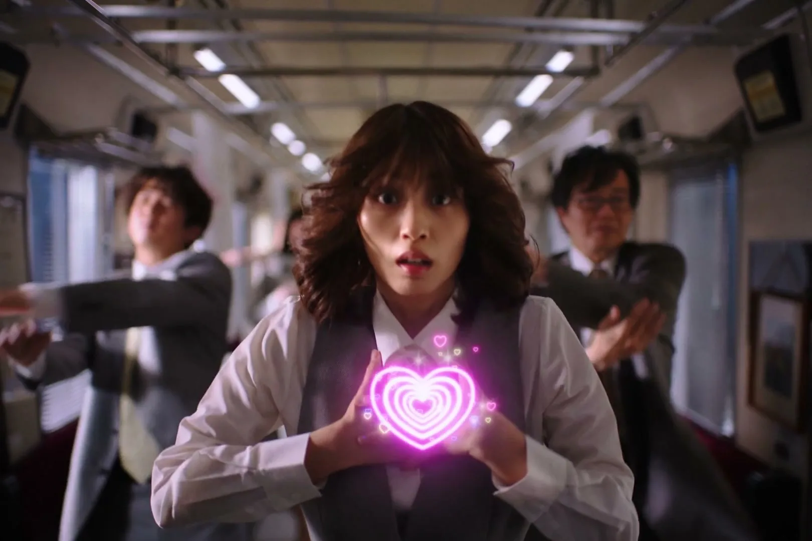 A young woman in a business suit holds a glowing pink heart in her hands, looking surprised, while men in suits stretch in a train car.