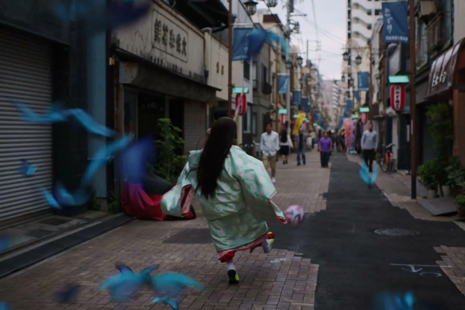 A young female in a traditional Japanese kimono runs down a narrow street, kicking a soccer ball, as blue butterflies flutter around her.