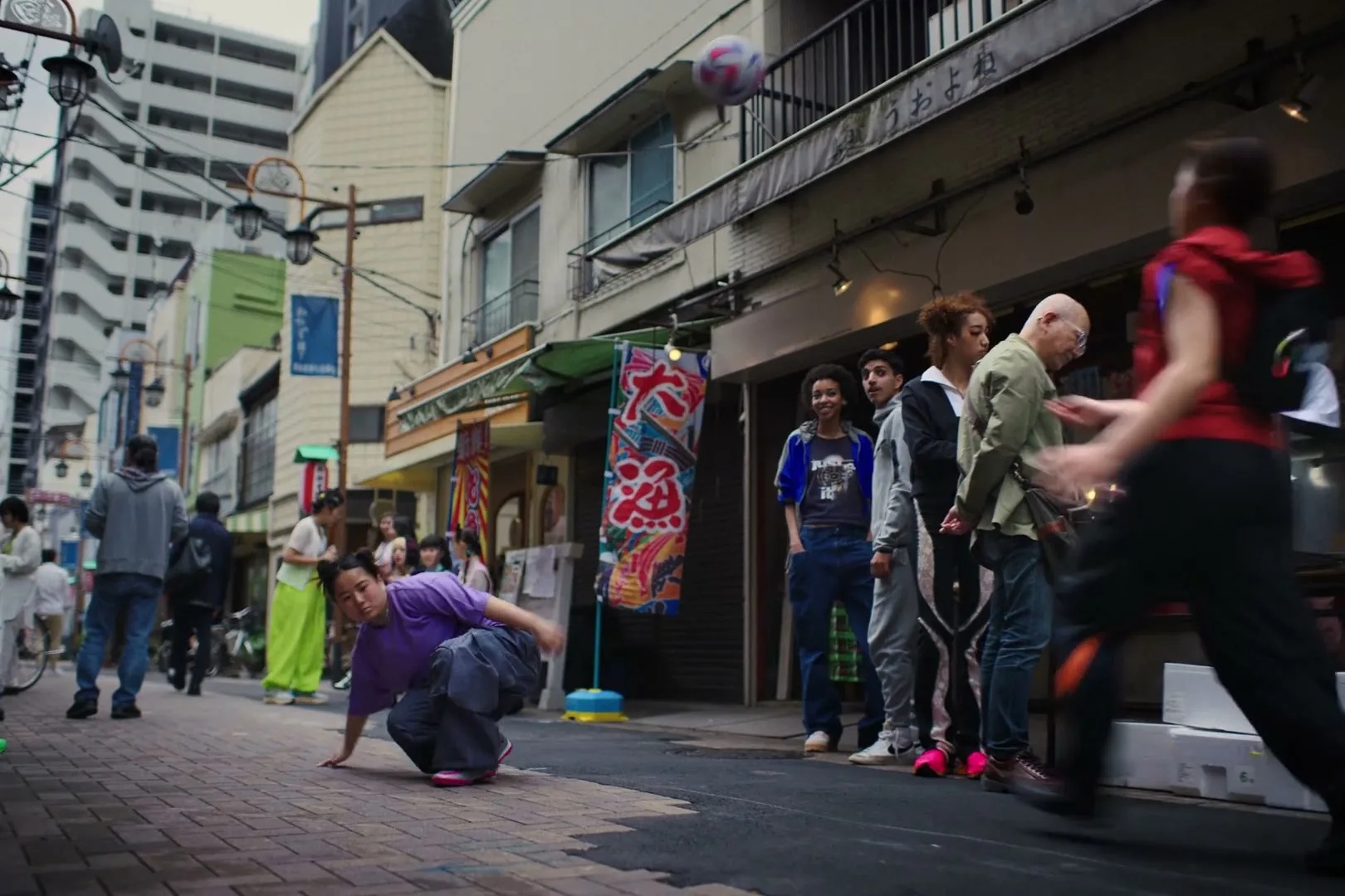 A young person in a purple shirt and dark pants performs a breakdancing move with a soccer ball on a brick sidewalk in a bustling city street.