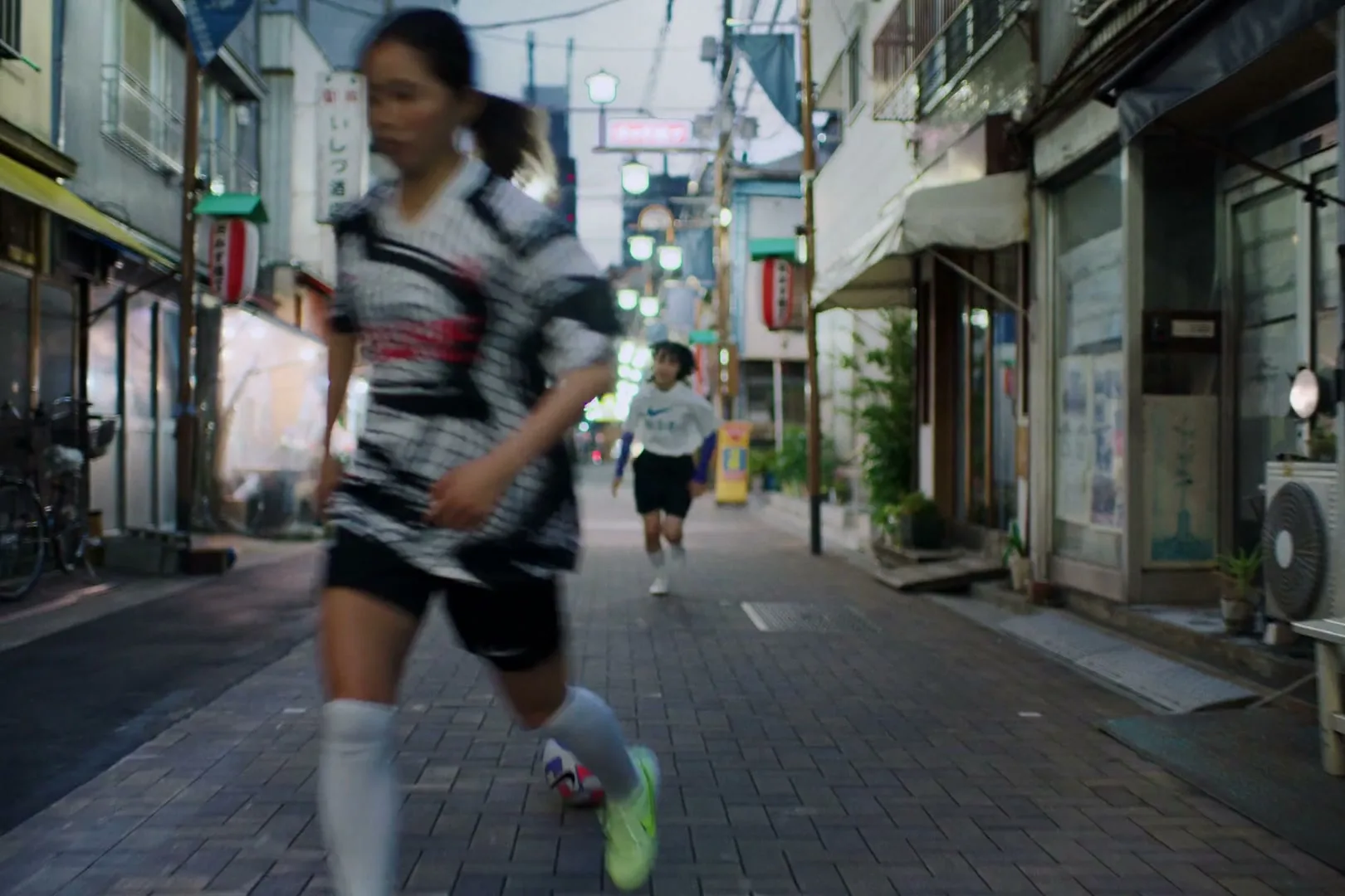 A young female soccer player in a black and white jersey dribbles a soccer ball down a narrow, illuminated street in a Japanese city at dusk.
