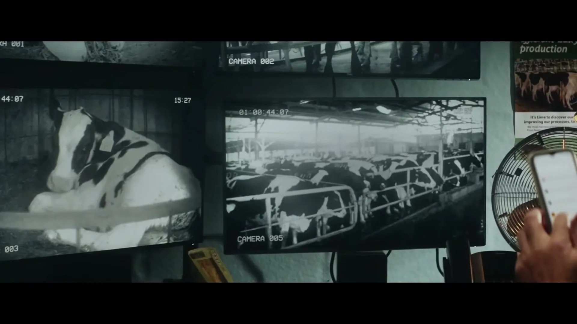 A person's hands hold a smartphone, scrolling through content, while multiple security camera feeds of cows in a barn are displayed on monitors in the background.