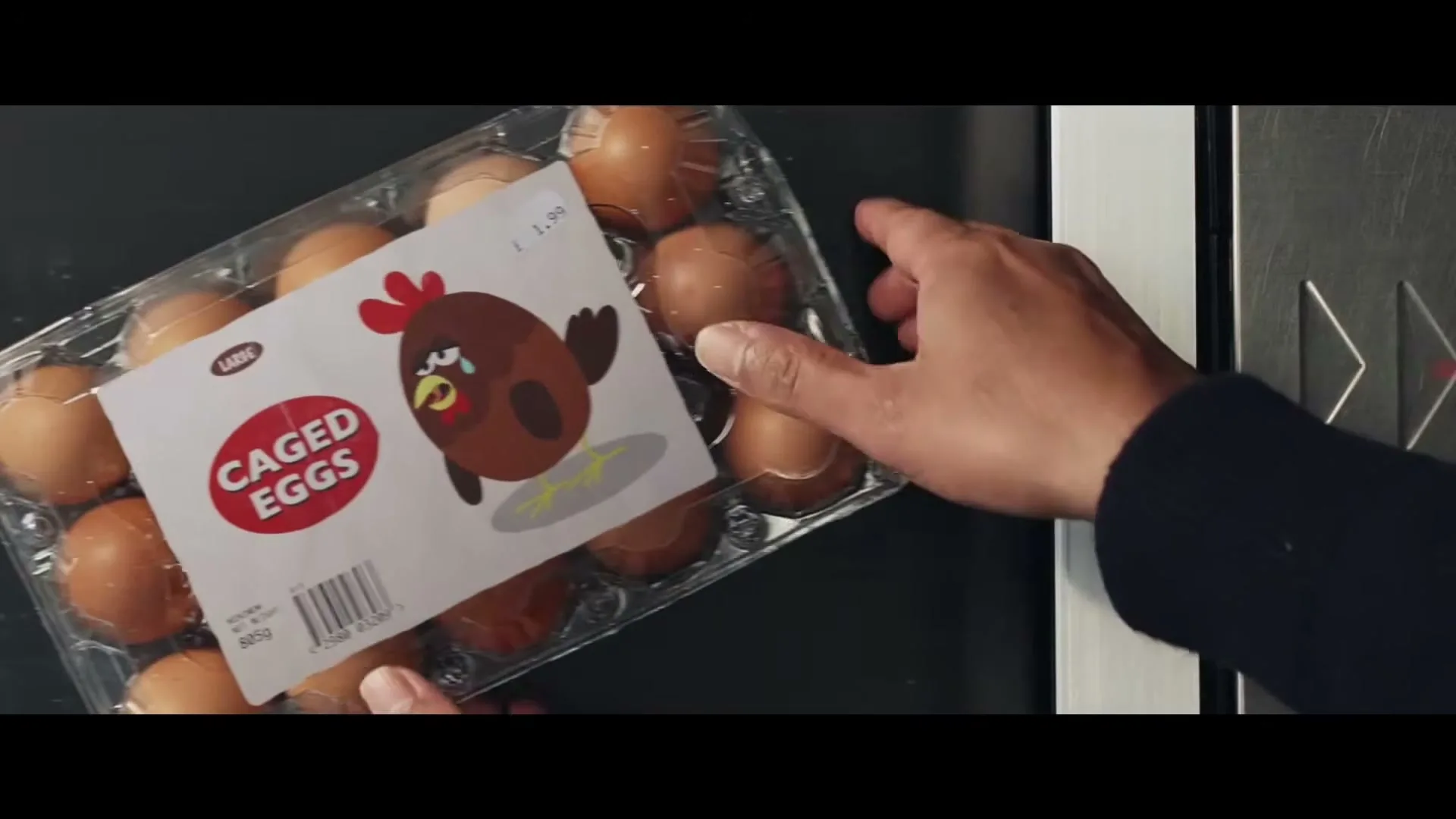 A person's hands place a carton of "Caged Eggs" with a sad chicken illustration onto a black surface next to an elevator button panel.