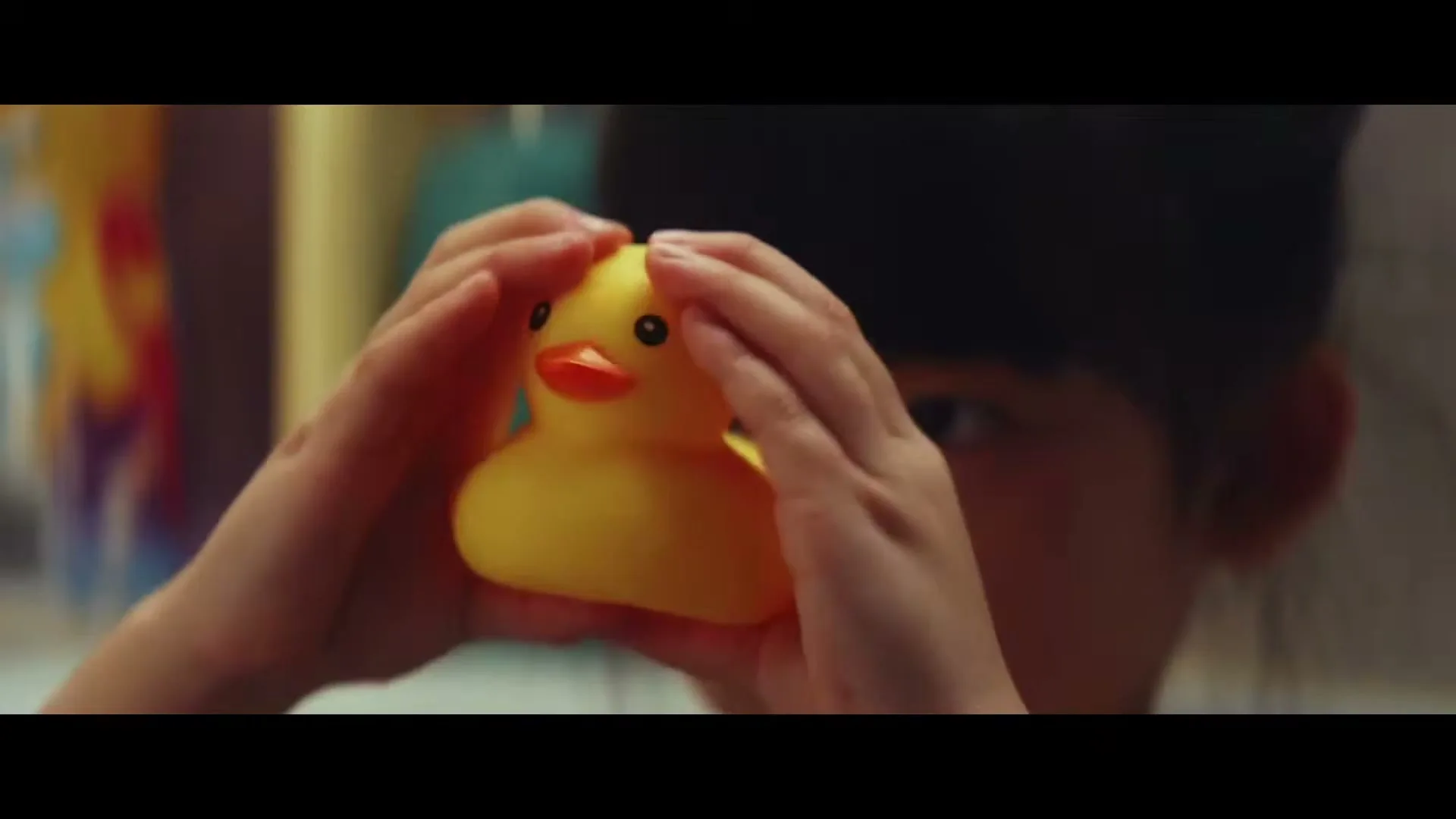 A child's hands hold a yellow rubber duck, with the child's face partially visible in the background, indoors.