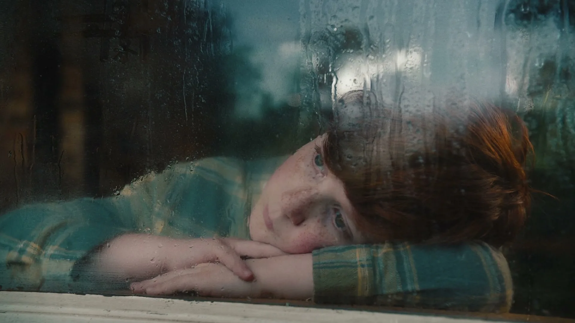 A young red-haired person with freckles rests their head on their arms, looking out a rainy window with a melancholic expression.