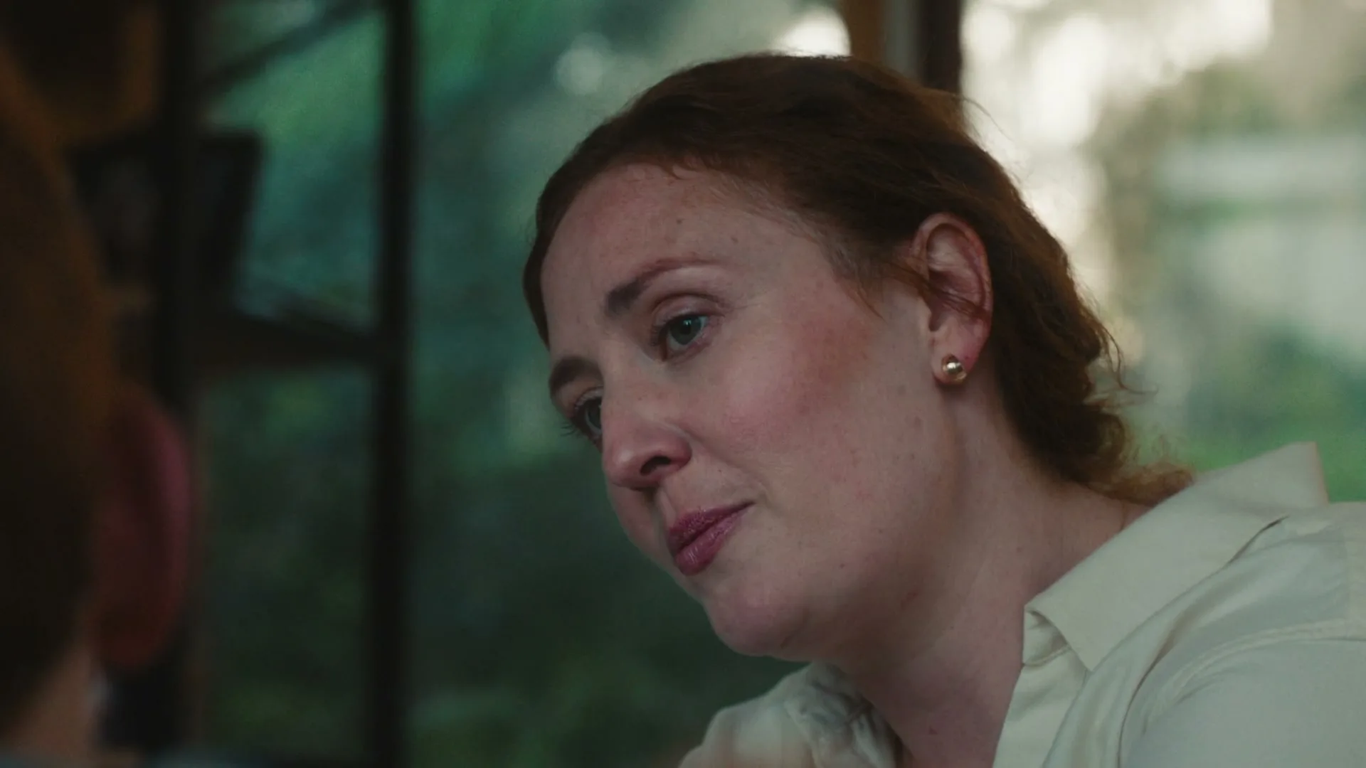 A woman with red hair and a white shirt looks down and to the left, smiling softly, in a warm, naturally lit interior setting.