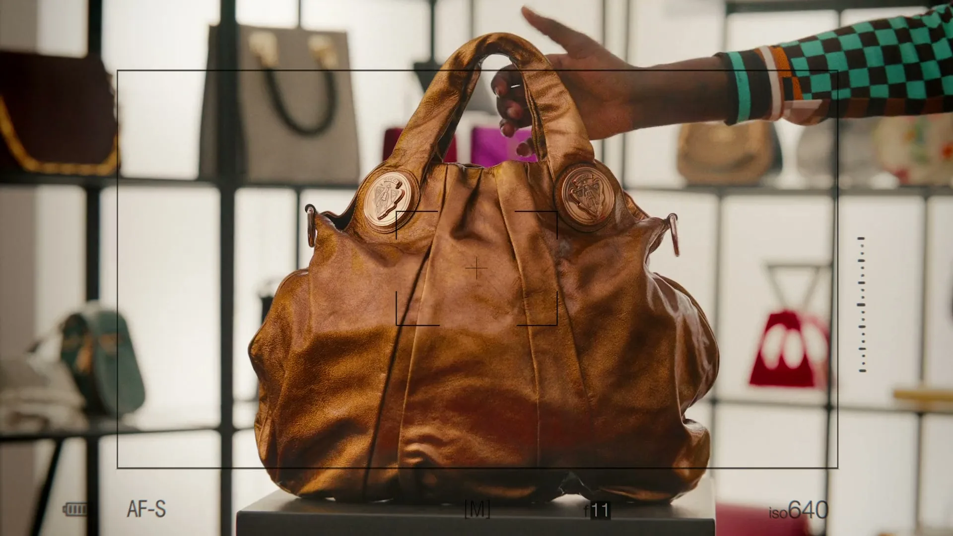 A dark-skinned hand places a brown leather handbag with gold accents onto a display stand in a brightly lit store, with other handbags visible in the background.
