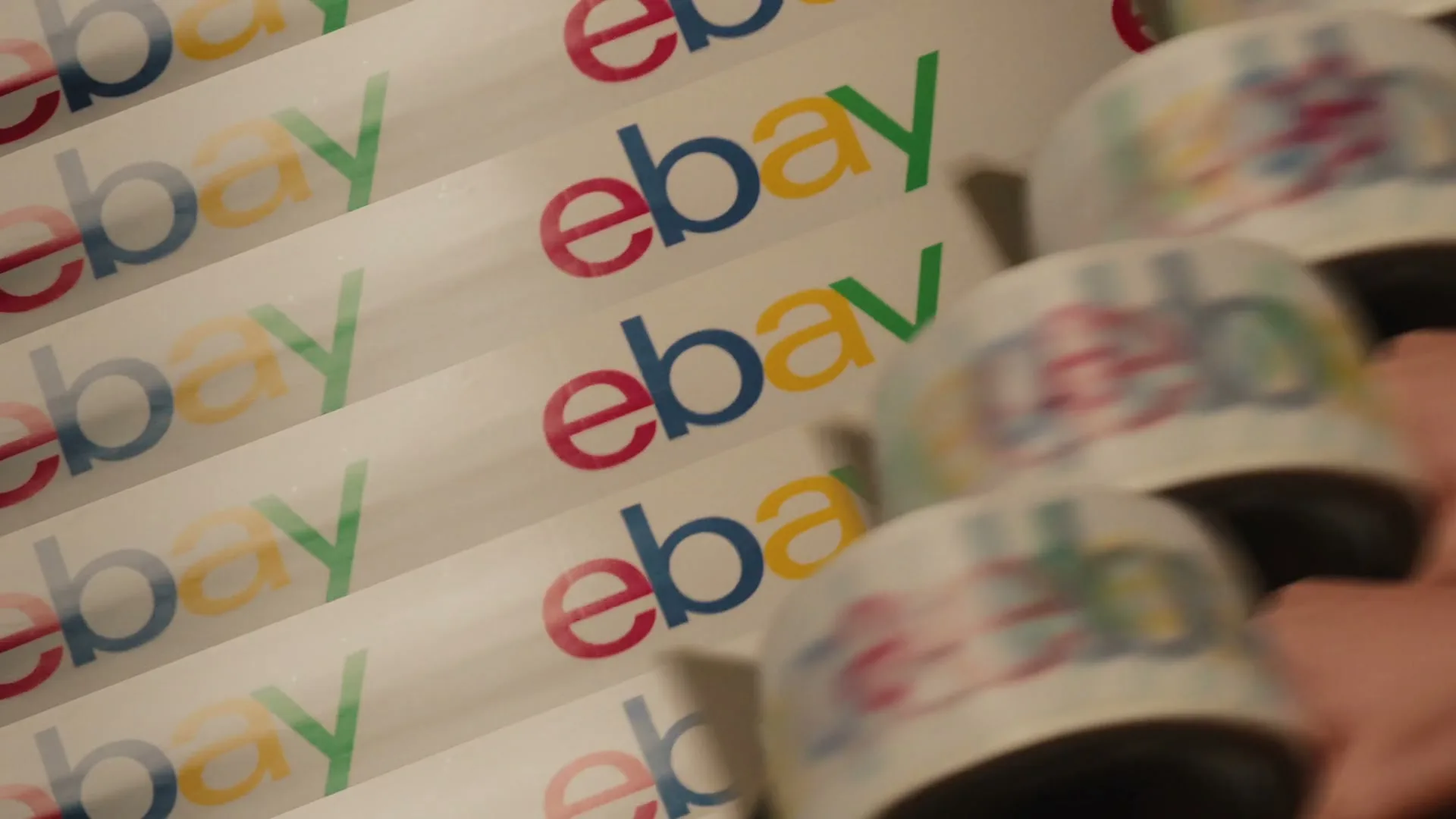Multiple hands are seen quickly placing white baseball caps with colorful logos onto a conveyor belt, which is also covered in colorful logos.