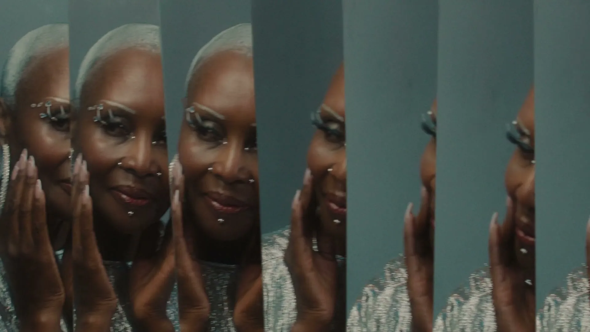 A woman with short white hair, wearing a sparkly top and large hoop earrings, smiles while looking at her reflection in multiple mirrors.