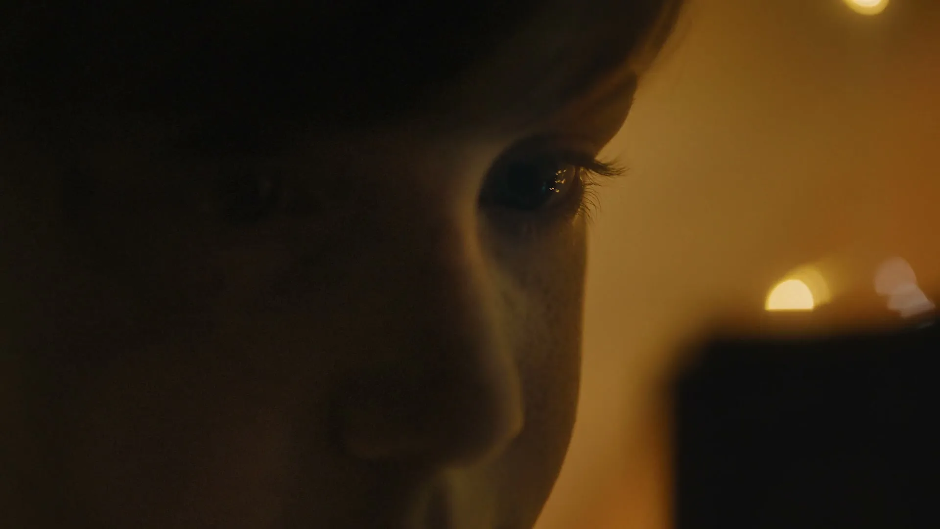 A child's face is seen in a dark room, illuminated by warm, out-of-focus lights in the background, creating a mysterious and serene atmosphere.