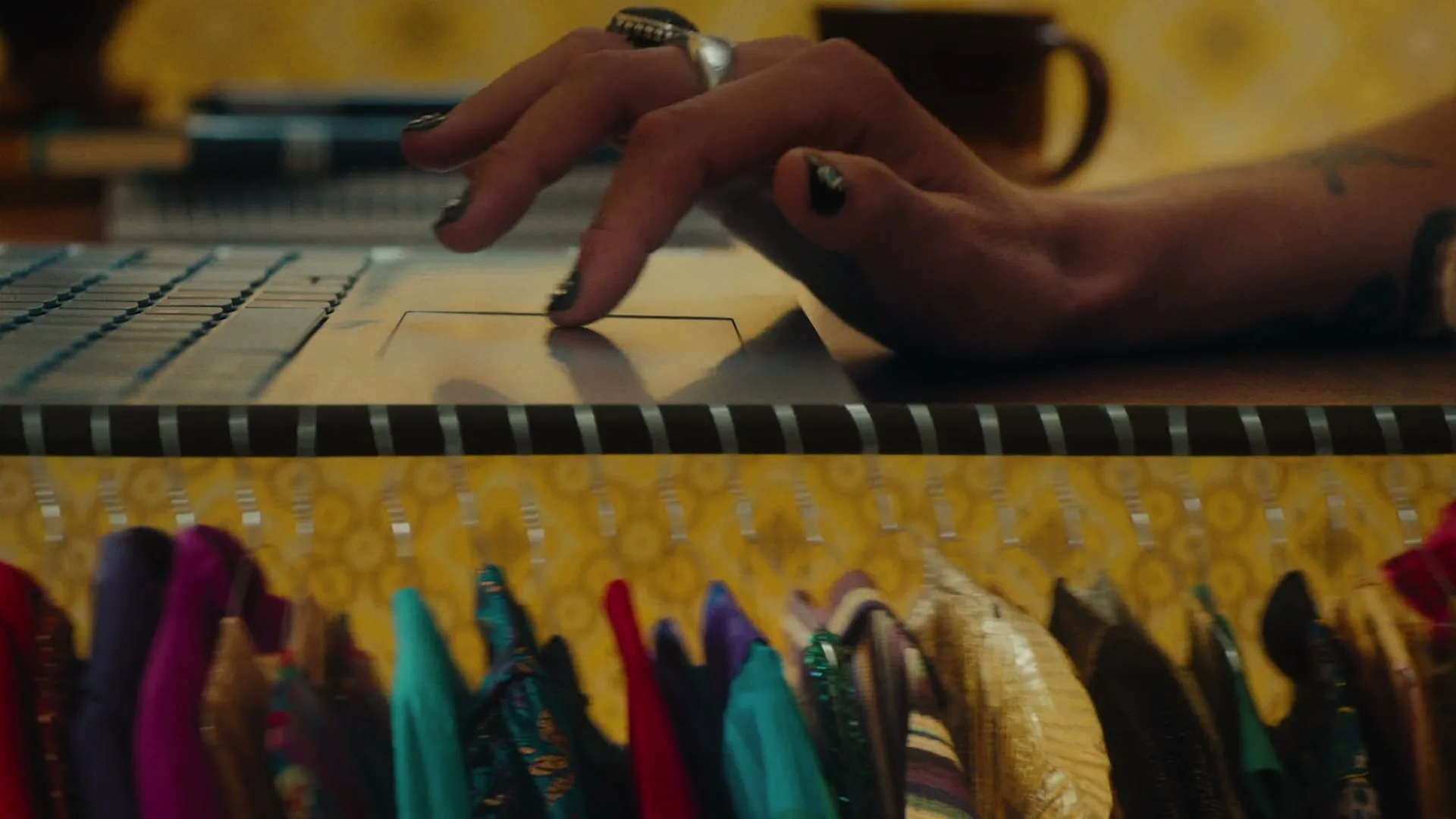 A person's hand with painted nails uses a laptop trackpad, while a rack of colorful clothes hangs below, in a cozy room.