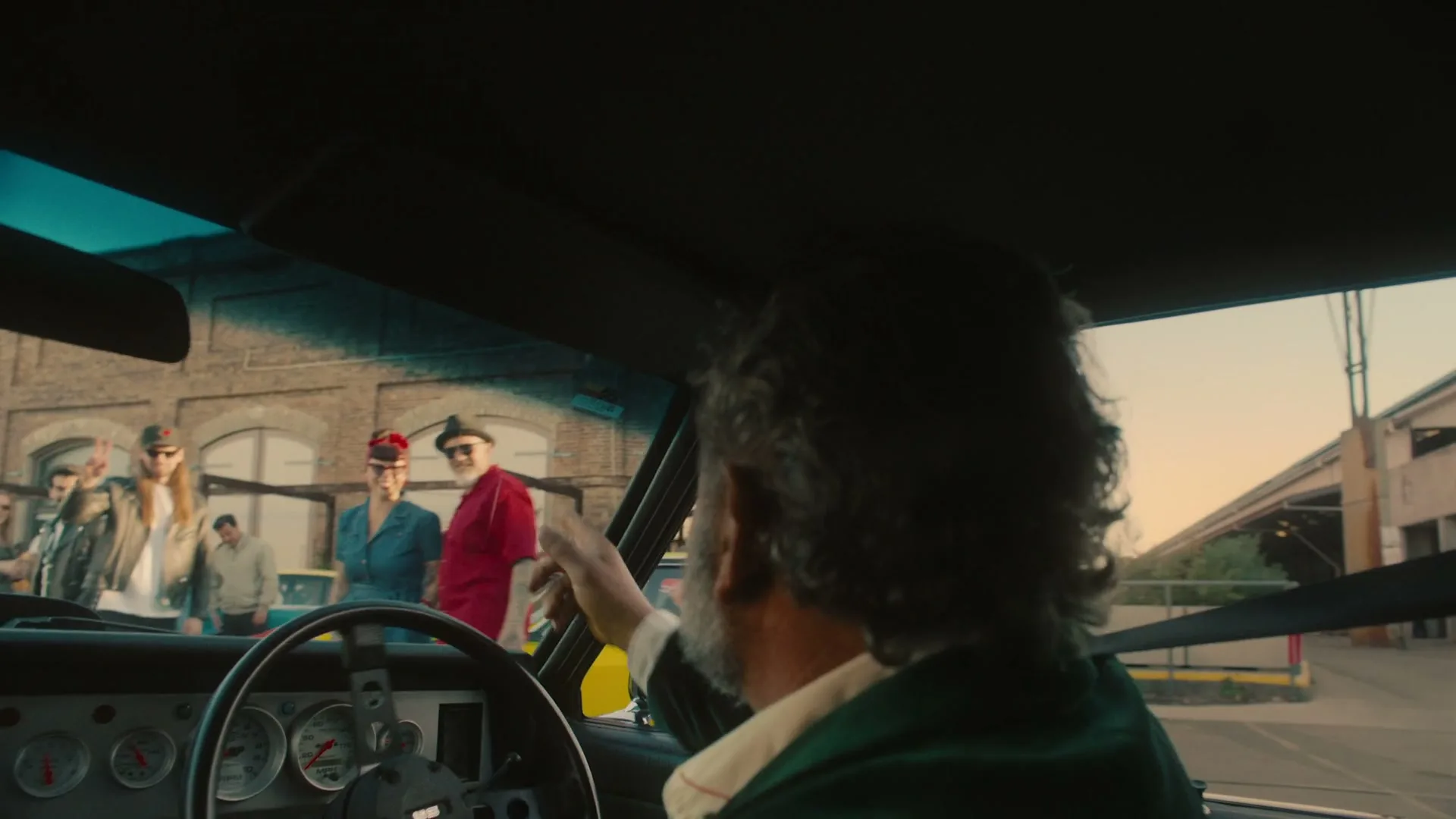 A man with a beard and curly hair, seen from behind, sits in a car and waves to a diverse group of people standing outside in an industrial area.