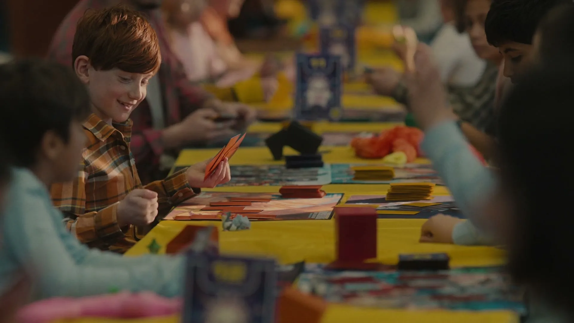 A red-haired boy in a plaid shirt smiles at the camera while playing a card game at a long yellow table with other children.