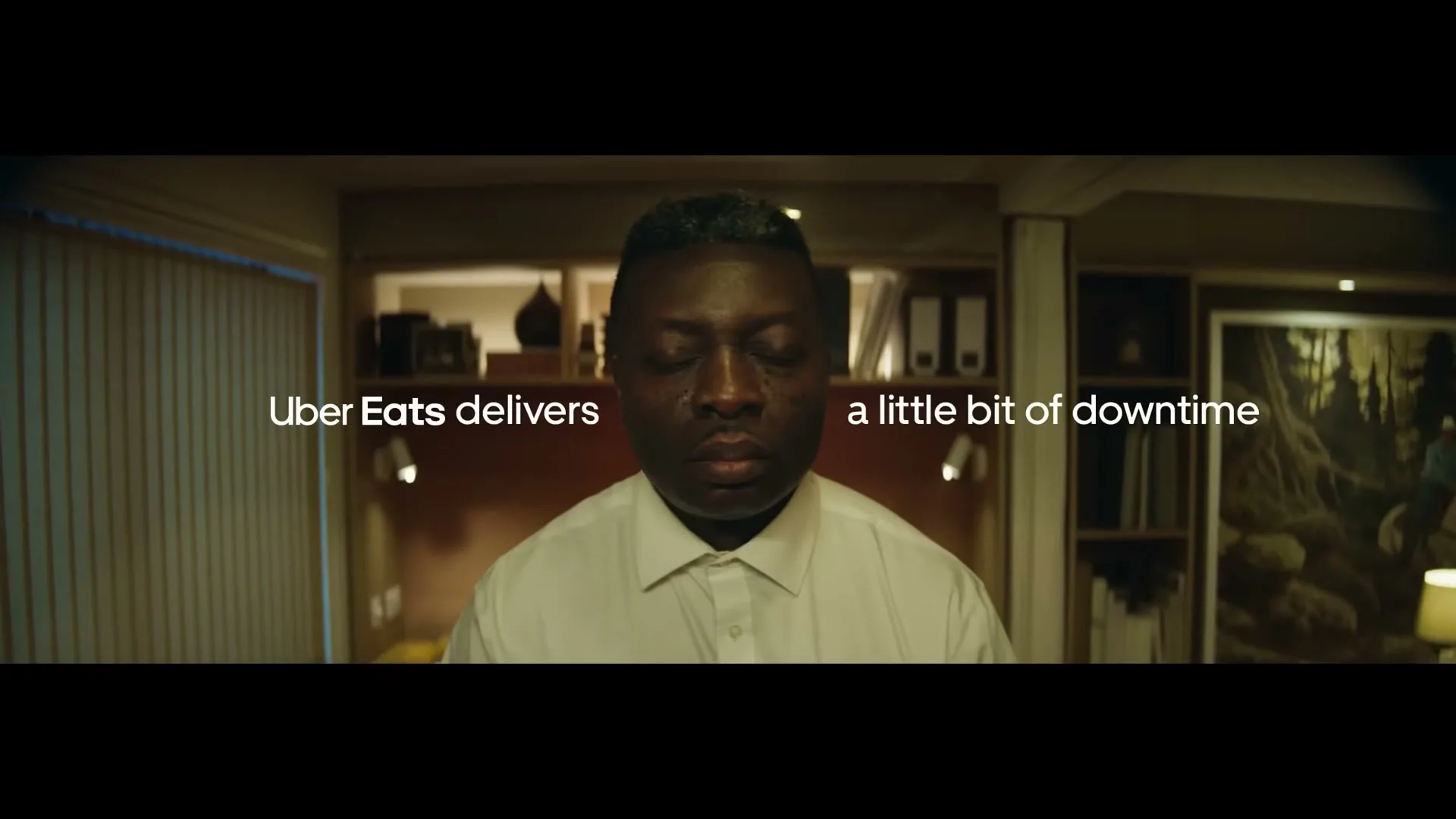 A Black man in his 30s, wearing a white shirt, looks at the camera, then looks down and to the right, in an office at night.
