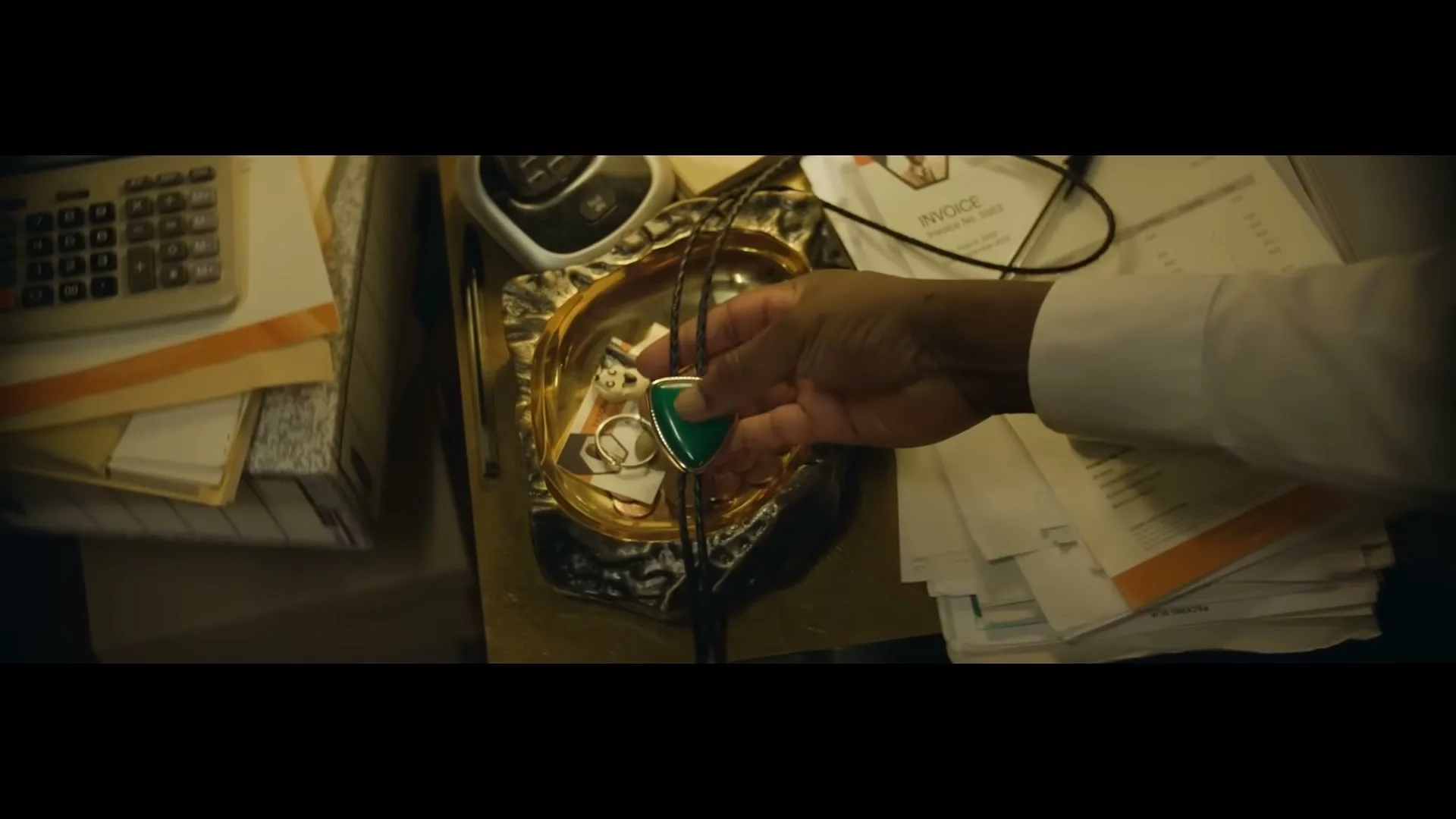 A man's hand picks up a green bolo tie from a decorative dish filled with coins and small items on a cluttered desk.