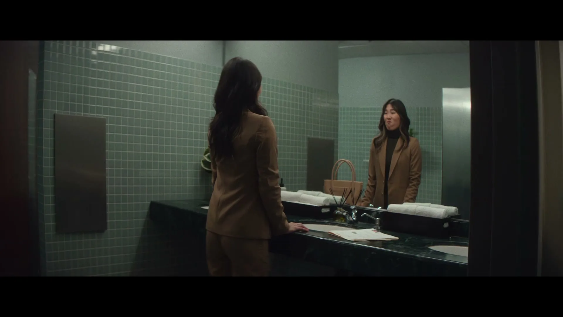 A woman in a brown suit stands in a public restroom, looking at her reflection in the mirror, as another person walks past the open door.
