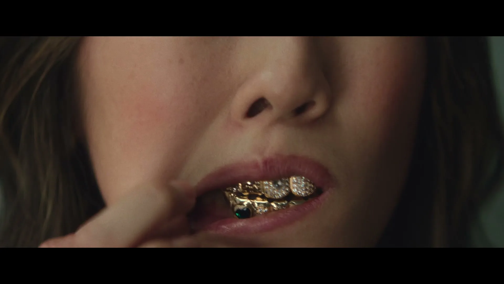 A woman with long brown hair and a pink top, wearing gold and diamond grills, touches her mouth with her finger in a closeup shot.