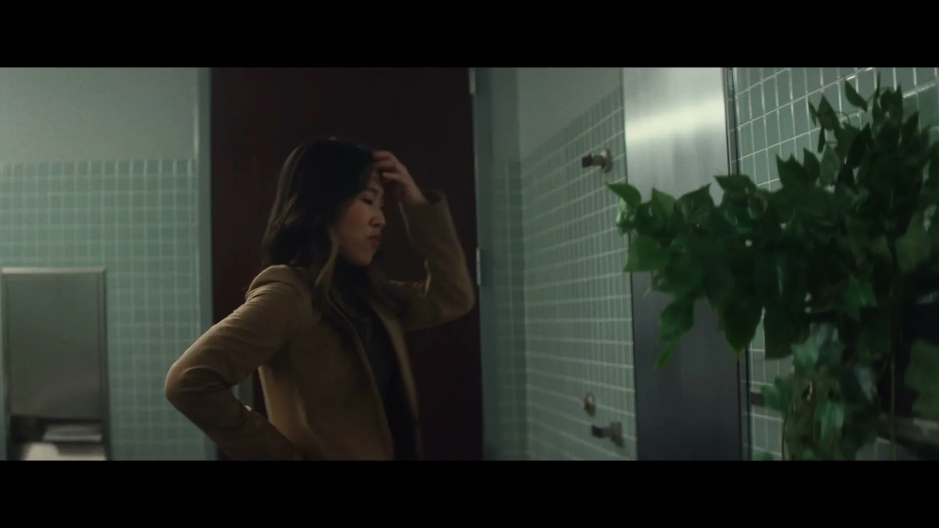 A young woman with long dark hair, wearing a brown jacket, stands in a tiled public restroom, looking frustrated and adjusting her hair.