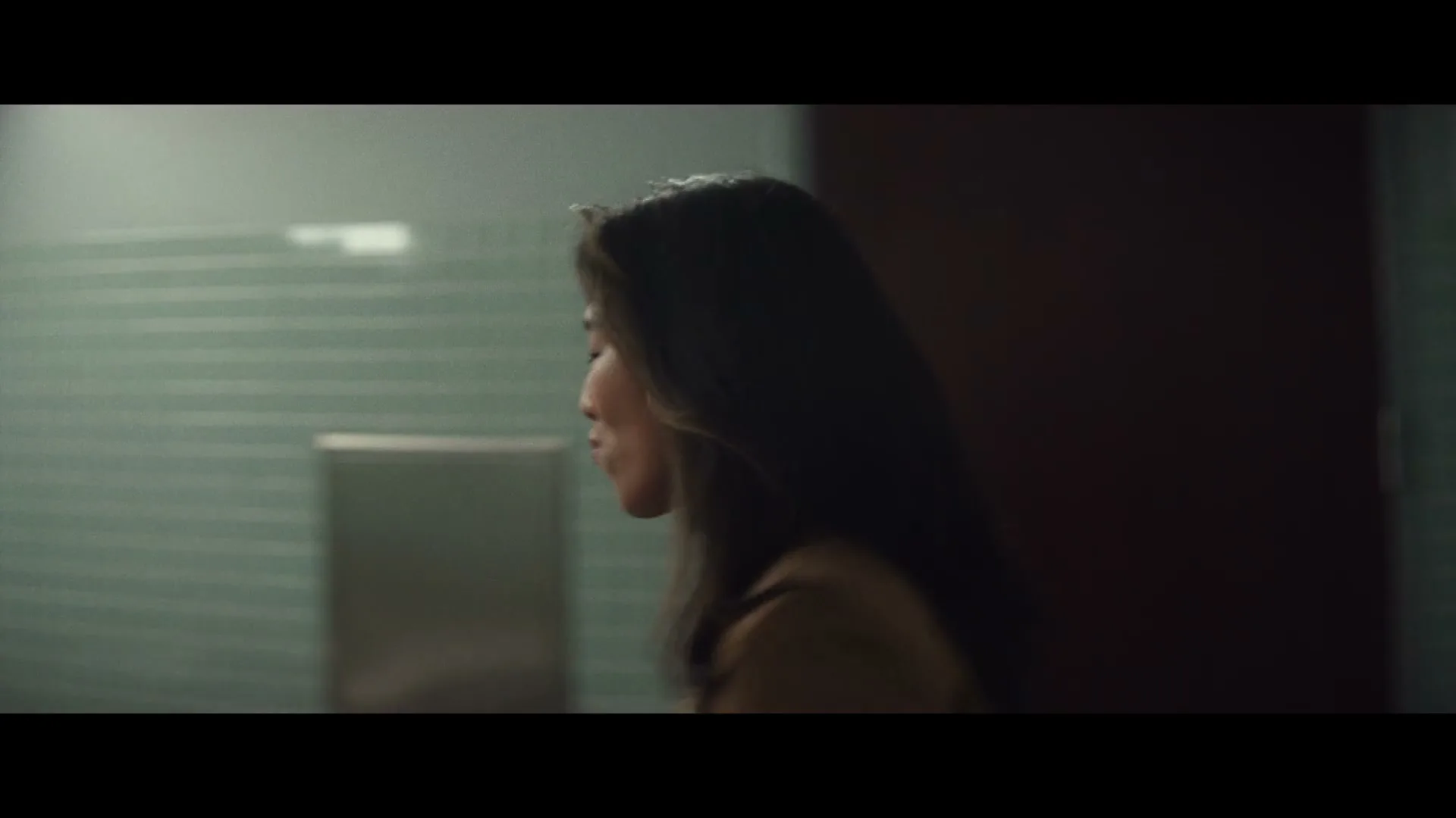A young Asian woman in a brown coat and black turtleneck looks at herself in a mirror in a public restroom.