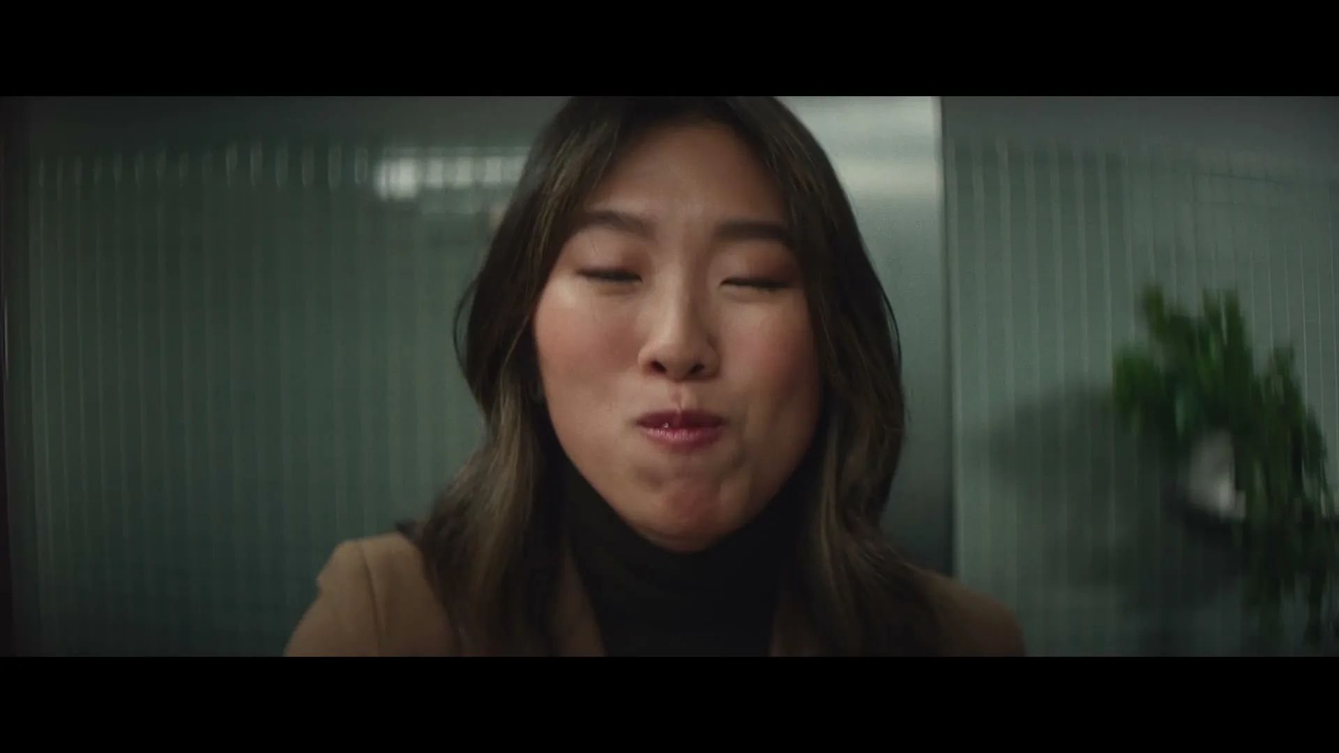 A young Asian woman with gold grills on her teeth smiles and talks to the camera in an office hallway.