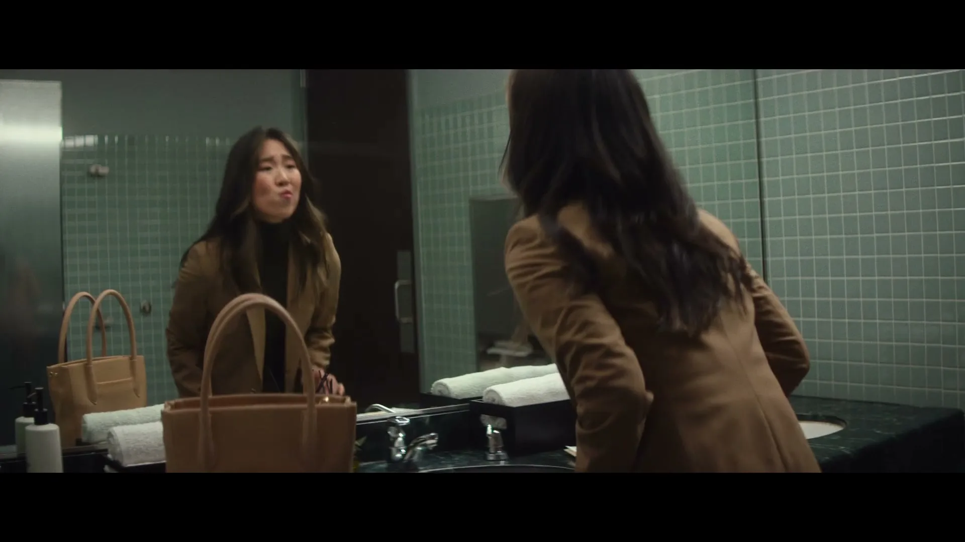 A young Asian woman in a brown blazer looks at her reflection in a public restroom mirror, making a funny face and picking at her teeth.