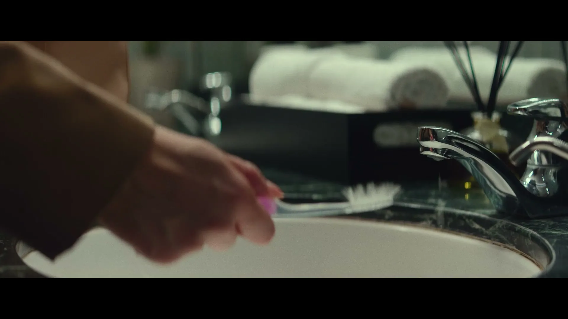 A person's hand holds a toothbrush over a bathroom sink with a dripping faucet, in a close-up shot.