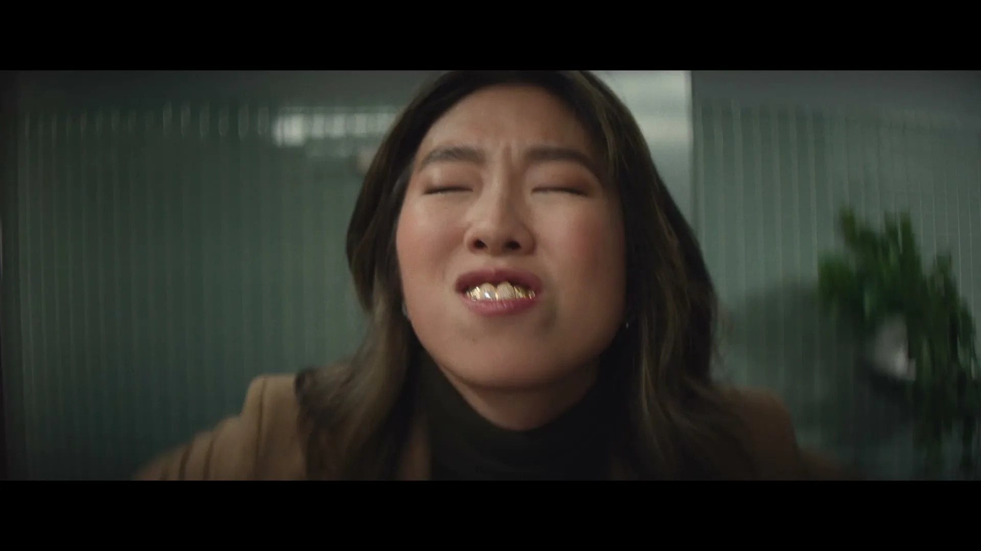 A young Asian woman with long dark hair and a brown jacket looks at the camera, then closes her eyes and reveals gold grills on her teeth, before turning away in a bathroom.