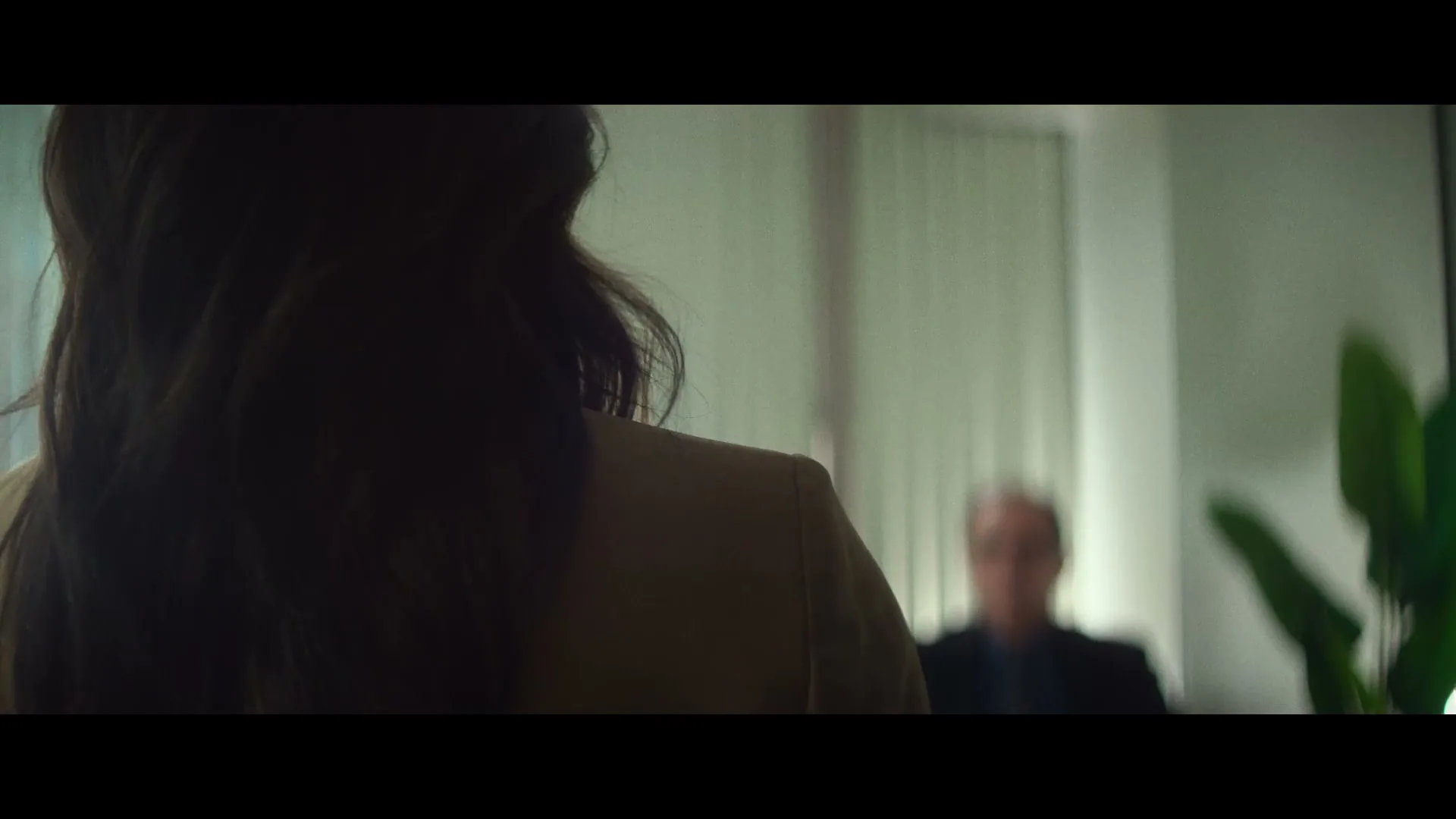 A woman with dark hair and a beige jacket is seen from behind, facing a blurry man in an office setting.