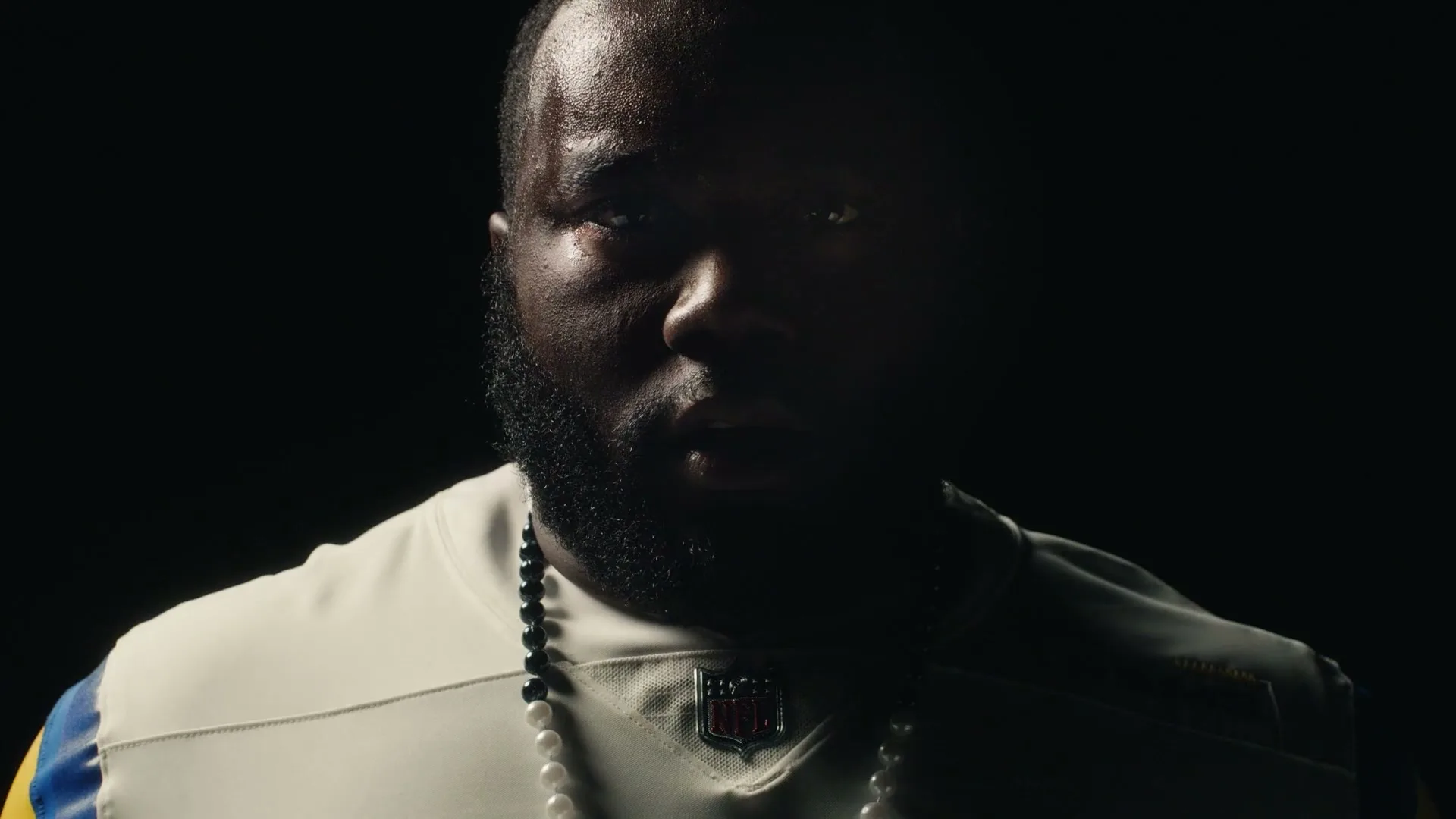 A Black man with a beard, wearing a white and blue football jersey and a pearl necklace, looks directly at the camera in a dark setting.