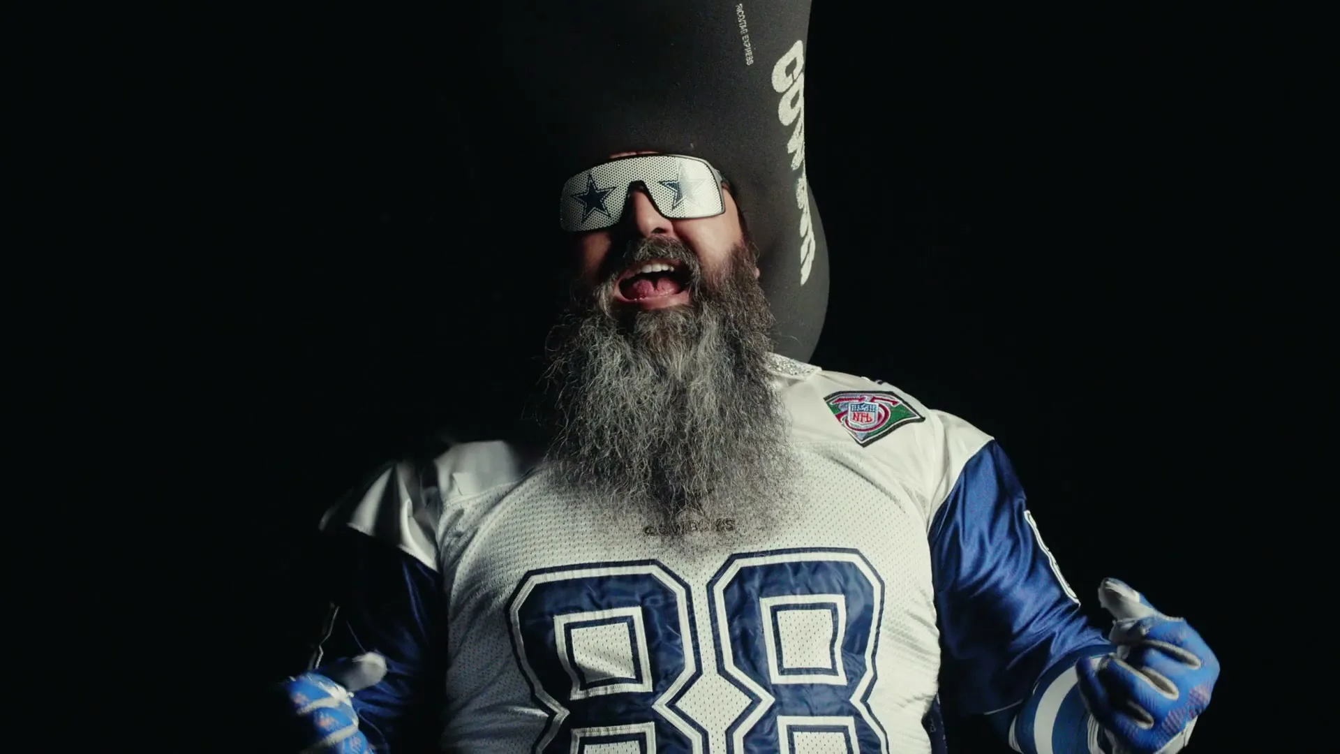 A man with a long grey beard, wearing a Dallas Cowboys jersey and star-shaped sunglasses, cheers enthusiastically in a dark void.