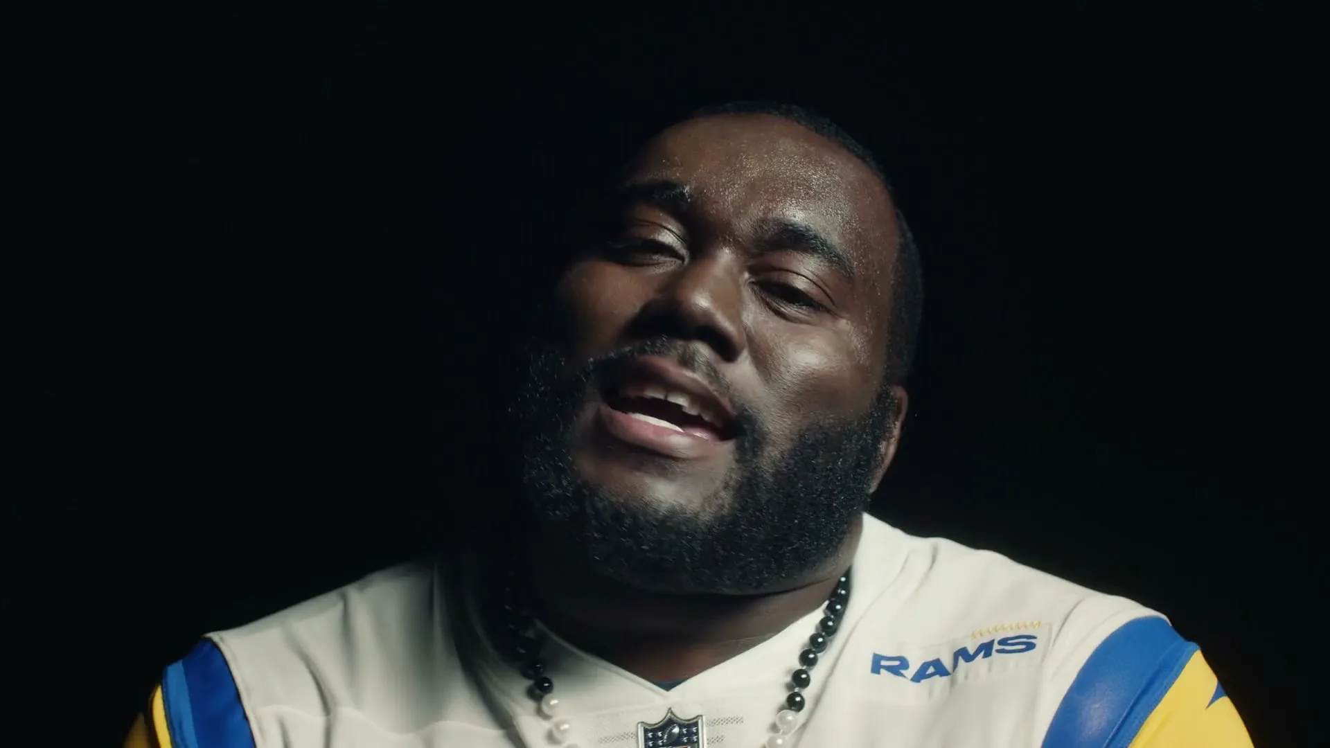 A man with a beard, wearing a white and blue football jersey and a pearl necklace, sings with his eyes closed against a dark background.