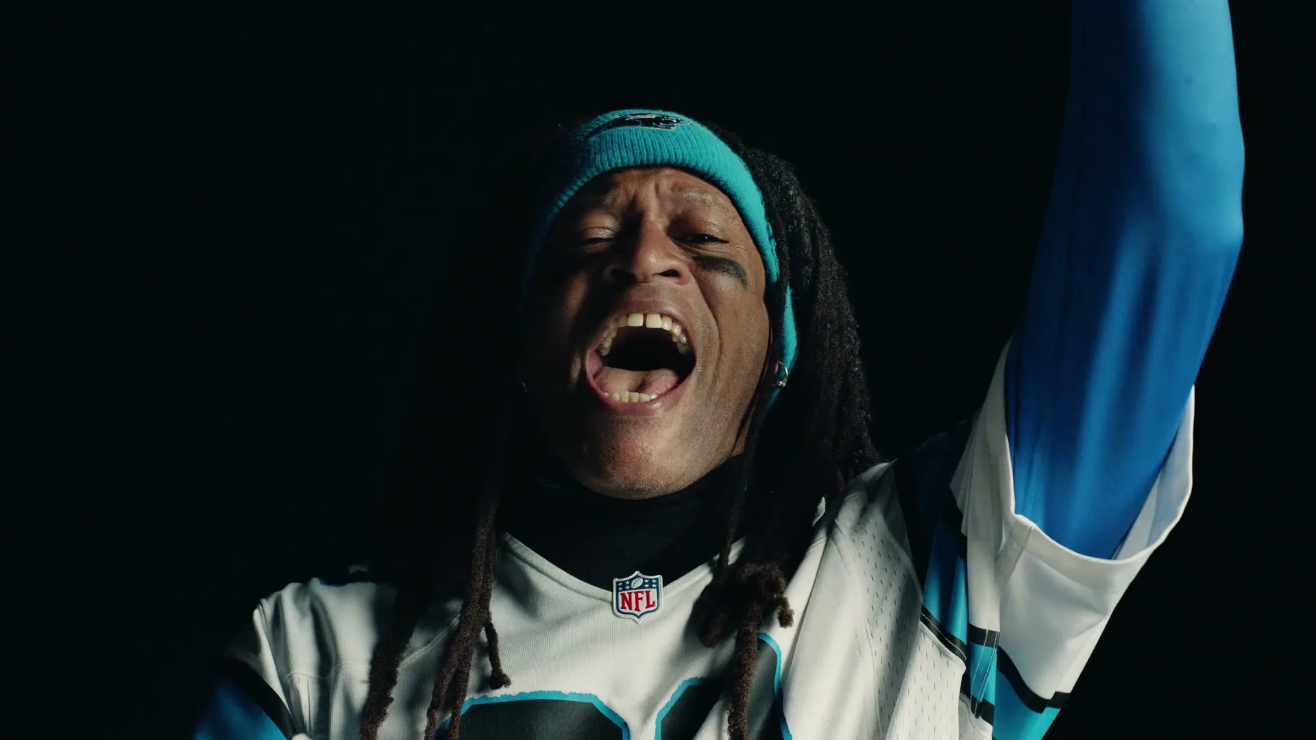A man with dreadlocks and a blue beanie, wearing a Carolina Panthers jersey, shouts with his arm raised in a dark setting.