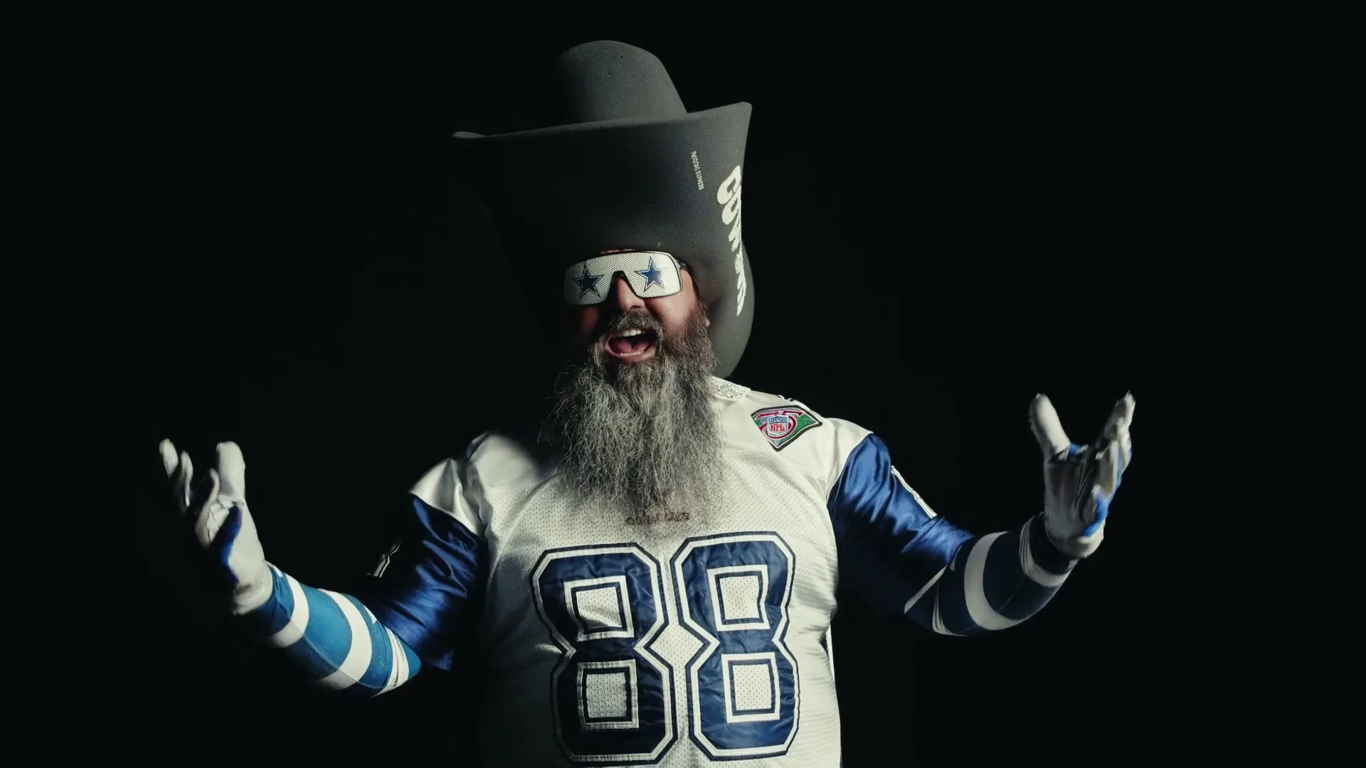 A man with a long grey beard, wearing a Dallas Cowboys jersey, a large cowboy hat, and star-shaped sunglasses, gestures excitedly in a dark studio.