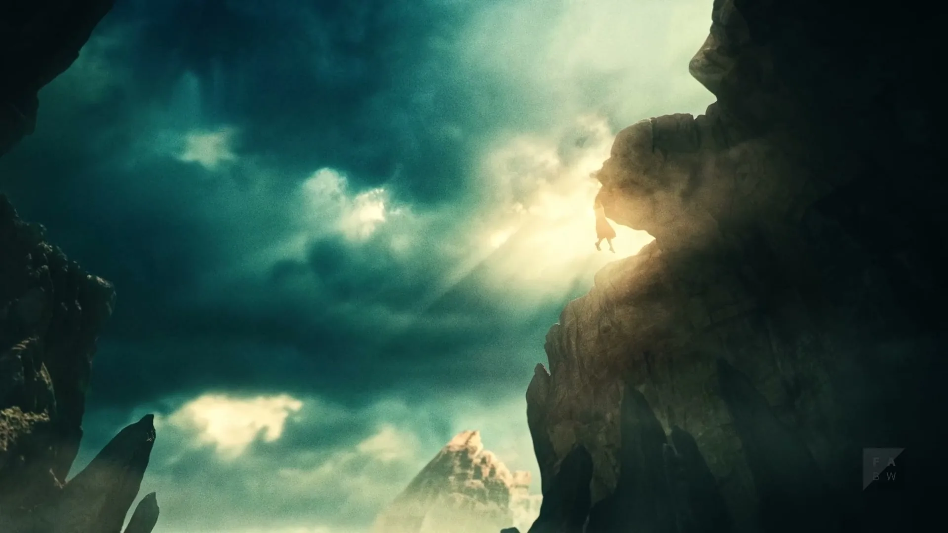 A small, silhouetted figure hangs precariously from a cliff edge, illuminated by dramatic sun rays breaking through dark, cloudy skies in a vast, mountainous landscape.