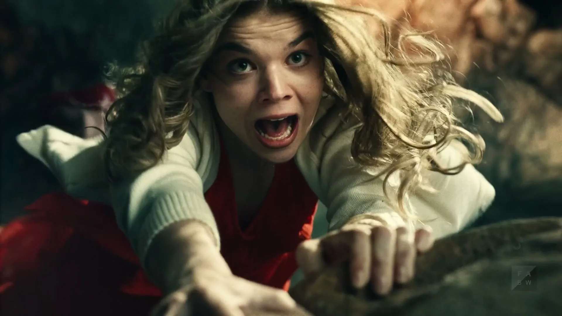 A young woman with blonde hair and a red dress struggles to climb up a rocky, dark surface, looking terrified.