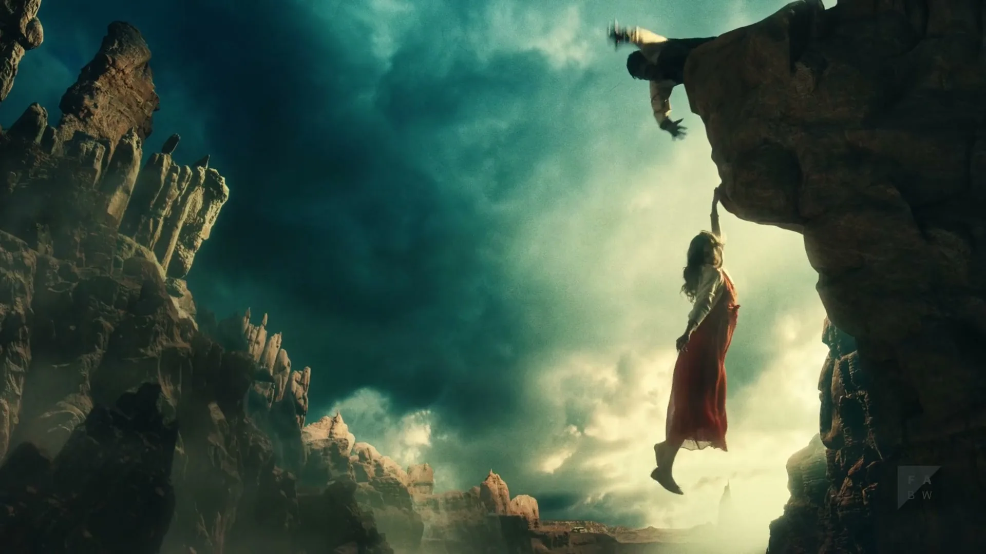 A man in a white shirt and dark pants holds onto a cliff edge, reaching for a woman in a red dress dangling below him, against a dramatic sky.