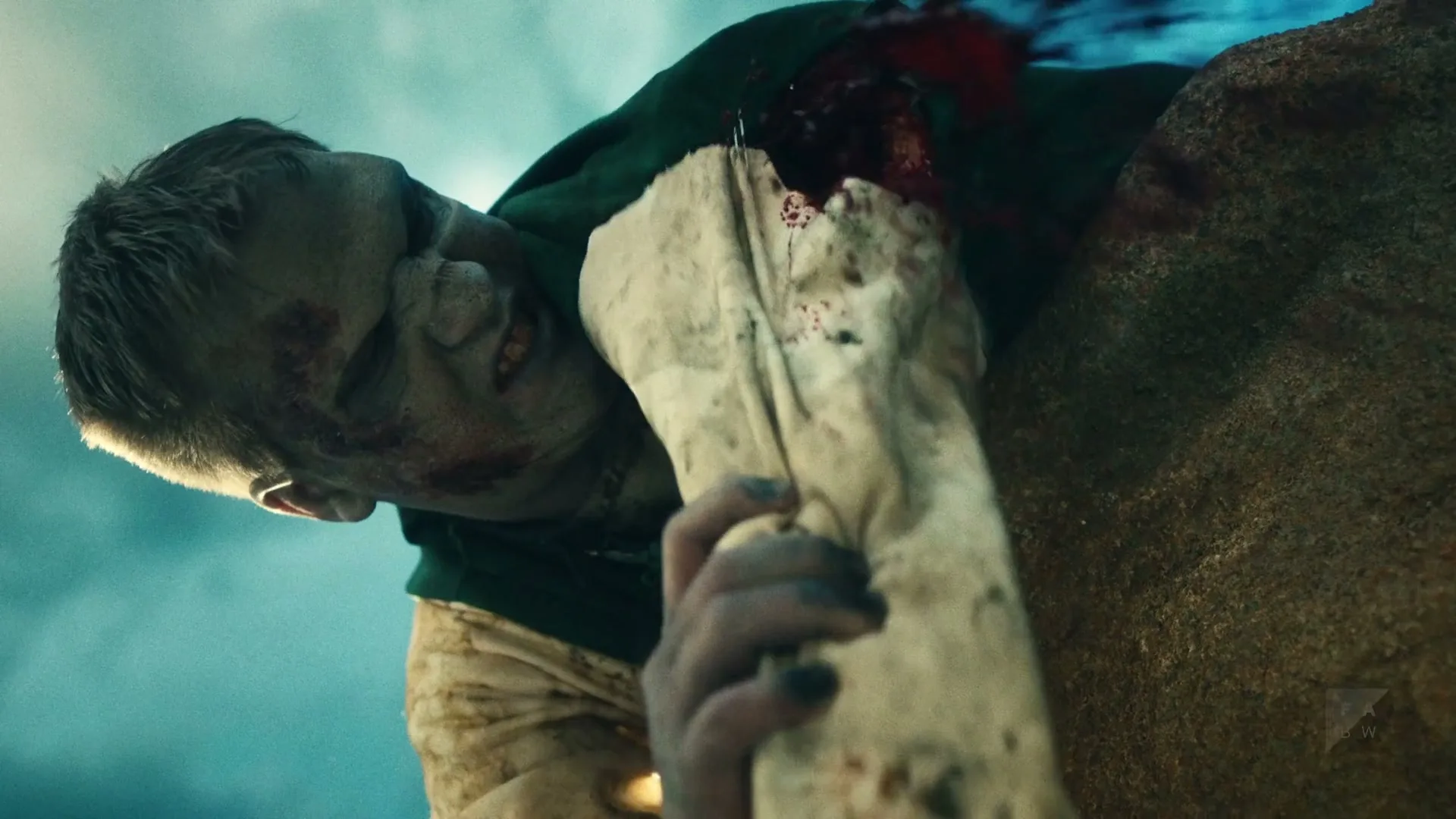 A zombie-like man with green skin and tattered clothes pulls a bone from his chest, looking pained against a blue sky.