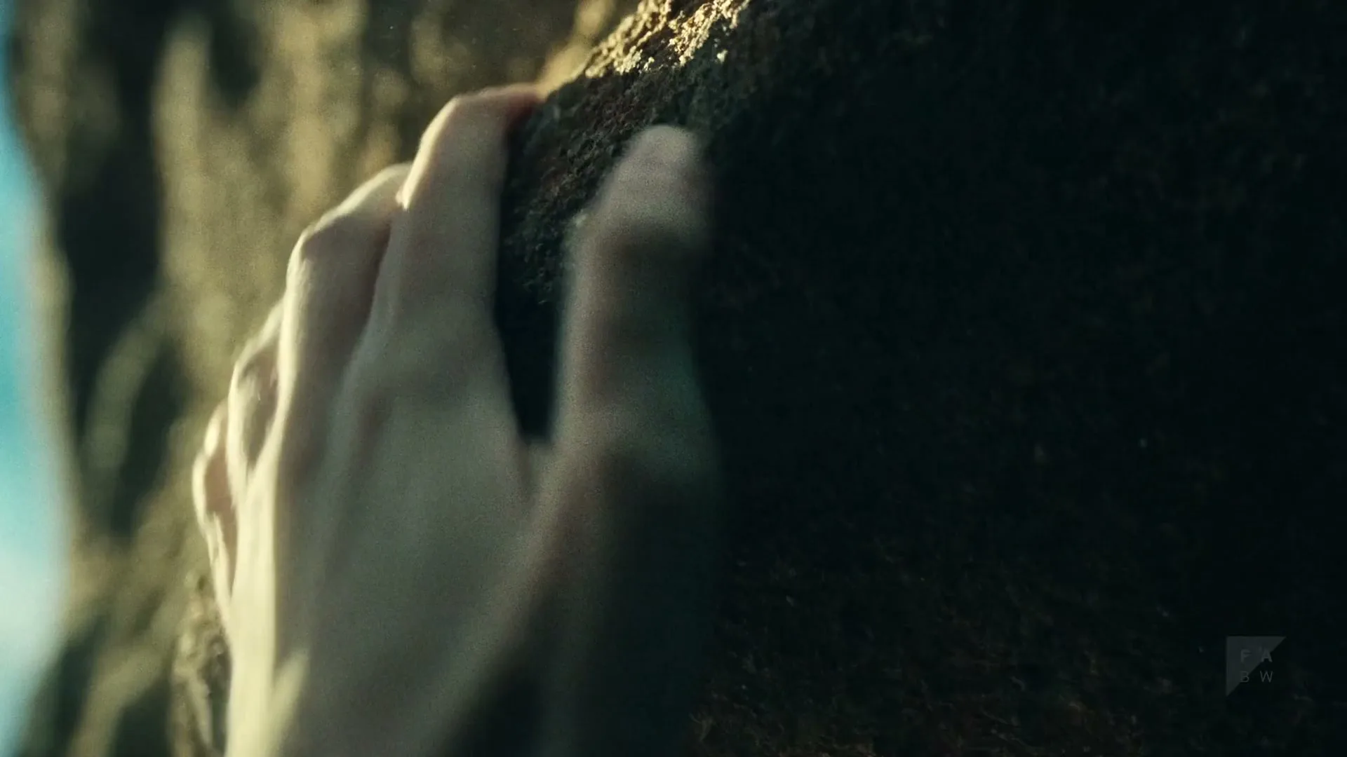 A pale hand with dirt under the fingernails grips a rough, dark rock, then slips, kicking up dust.