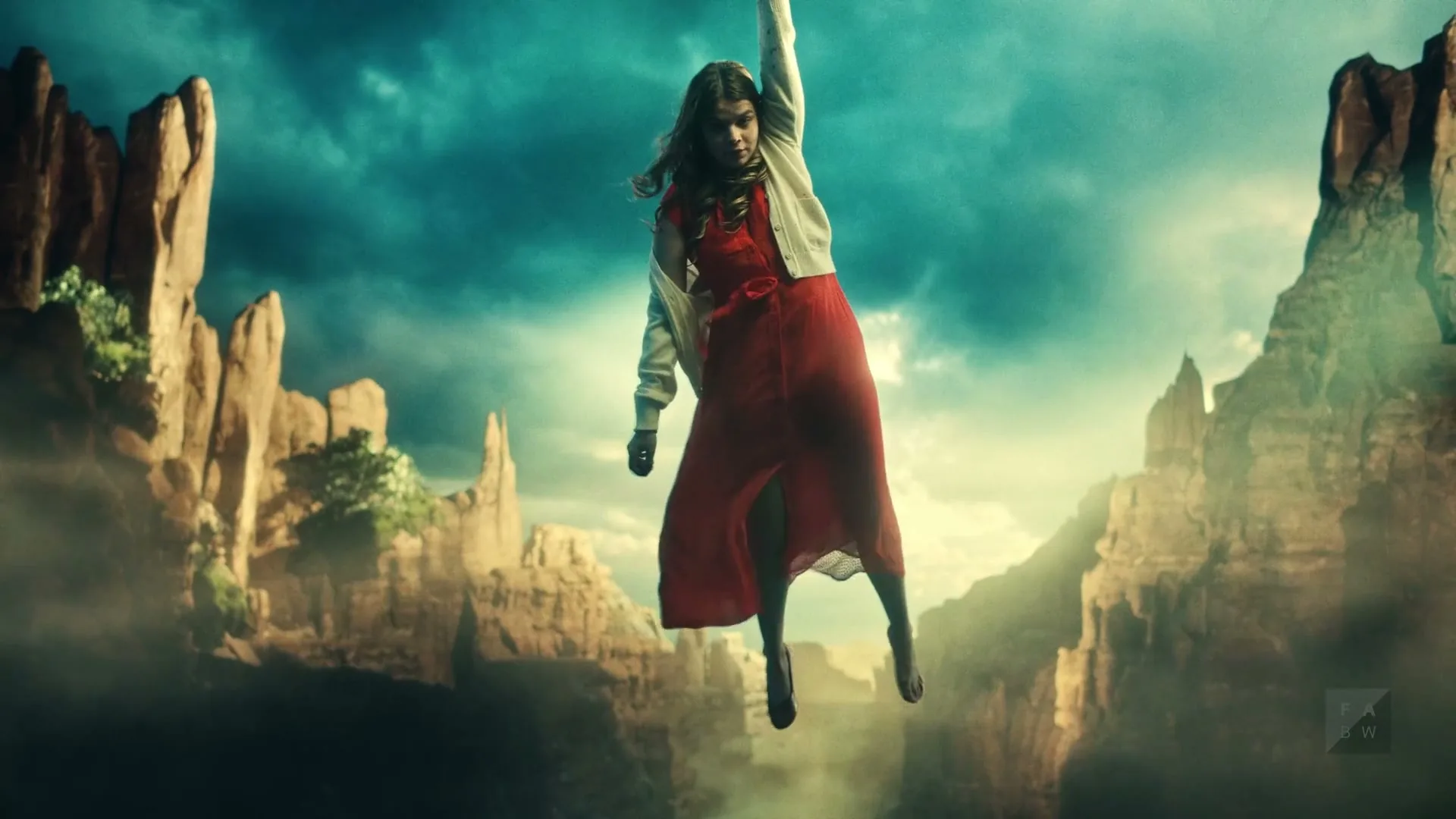 A young woman in a red dress and white cardigan floats in the air, suspended by an unseen force, against a backdrop of dramatic canyons and a cloudy sky.