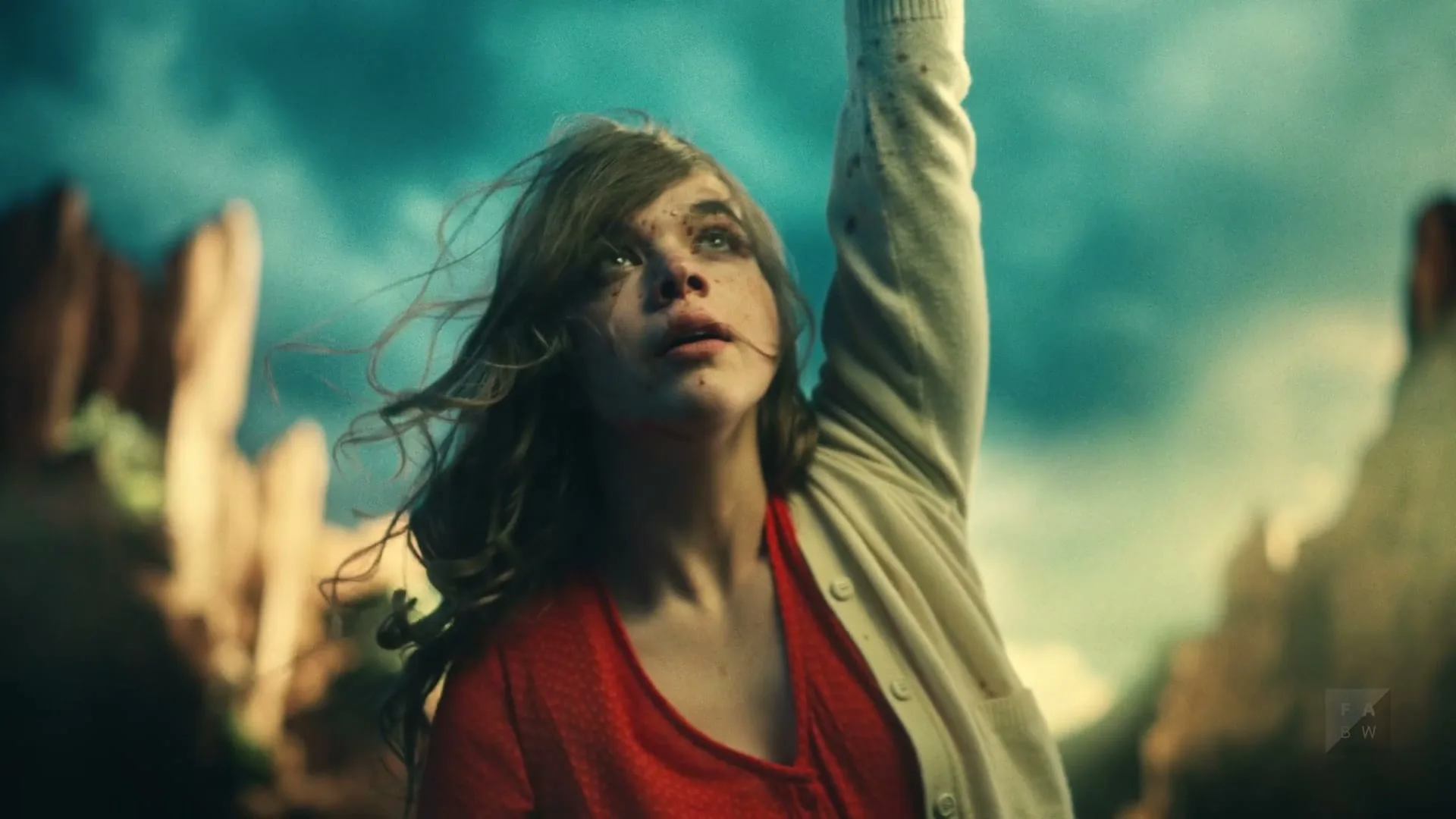 A young woman with dirty face and messy hair, wearing a red shirt and white cardigan, reaches her arm up towards a dramatic sky in a desert landscape.