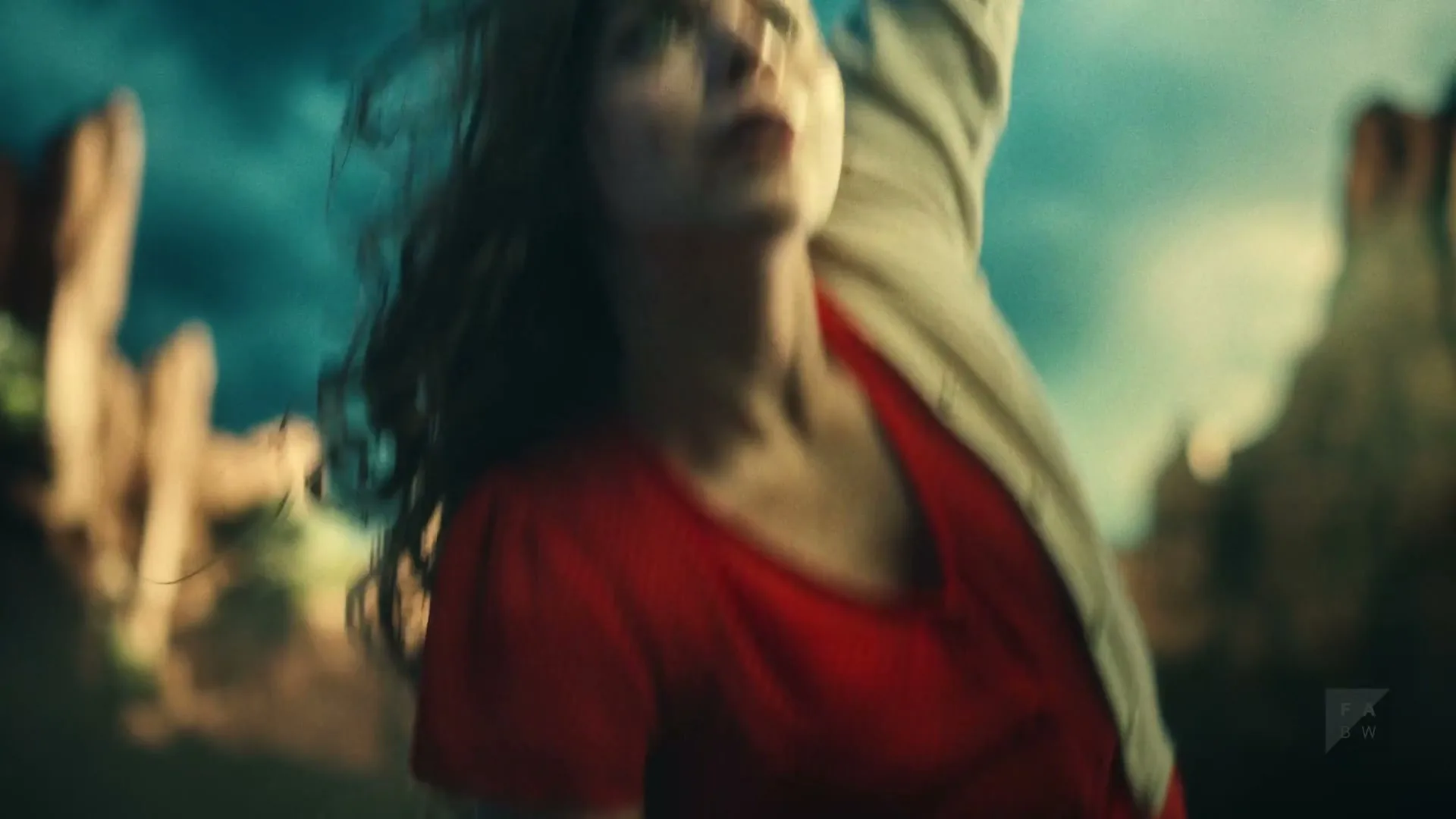 A young woman with dirty face and messy hair, wearing a red shirt and white cardigan, reaches her arm up towards a dramatic sky in a desert landscape.