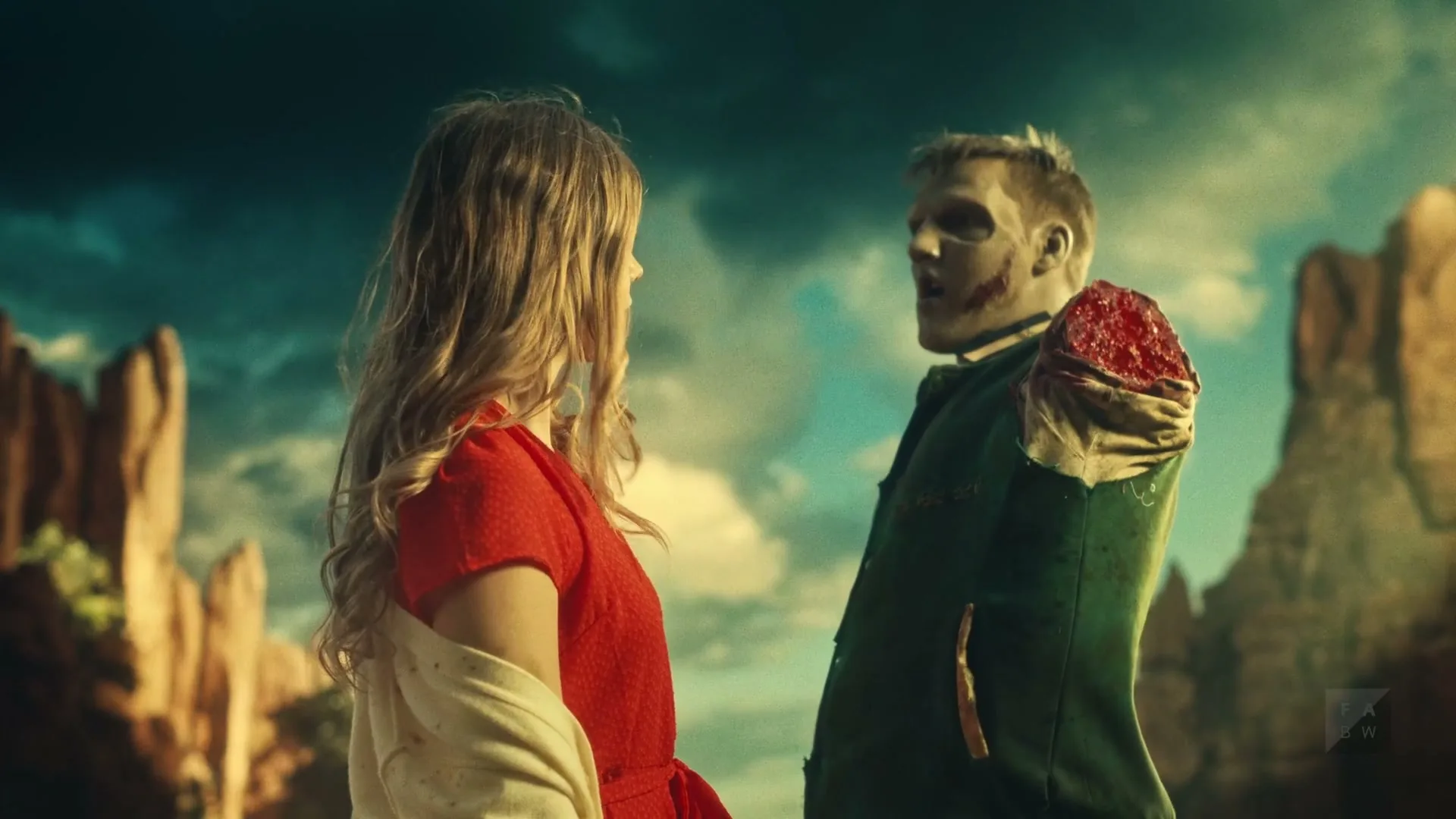 A young woman in a red dress and a zombie man with a missing arm look at each other, then the woman turns and walks away in a desert landscape.