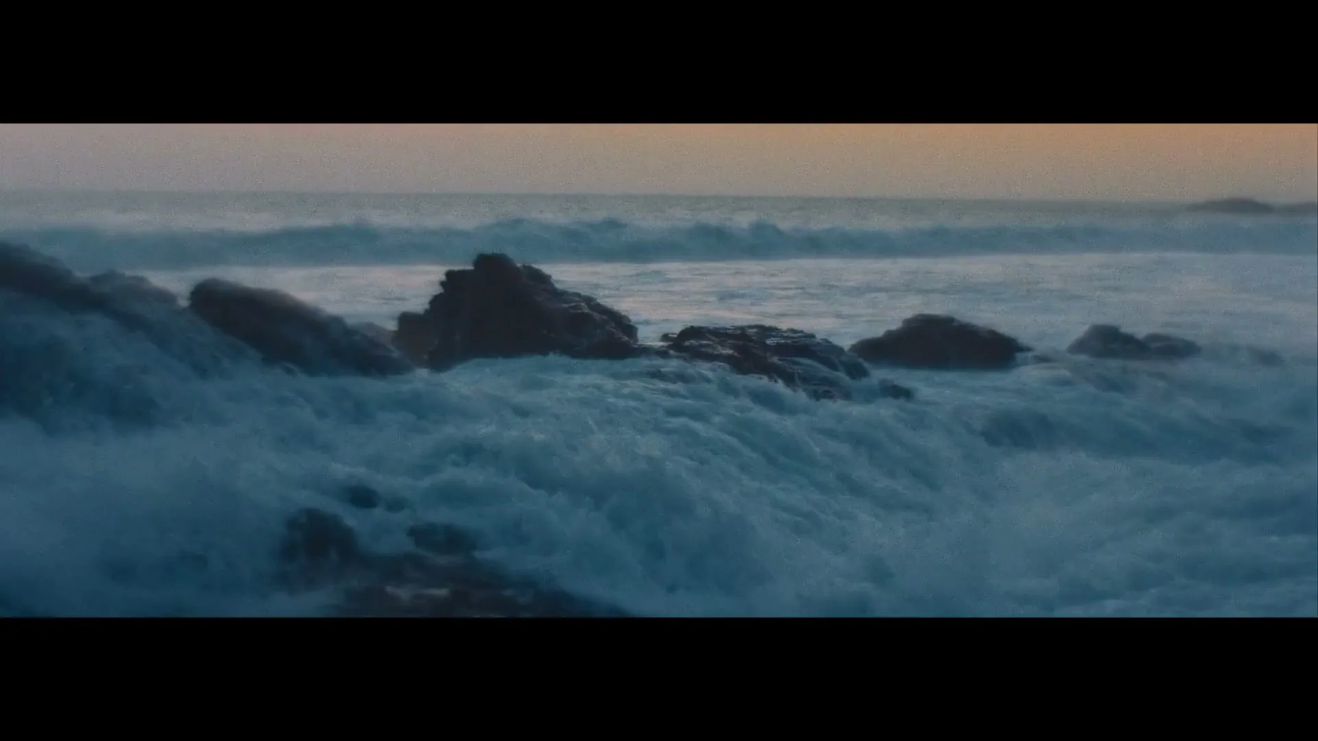 Waves crash against dark rocks on a stormy, overcast day at the ocean, with the horizon visible in the distance.