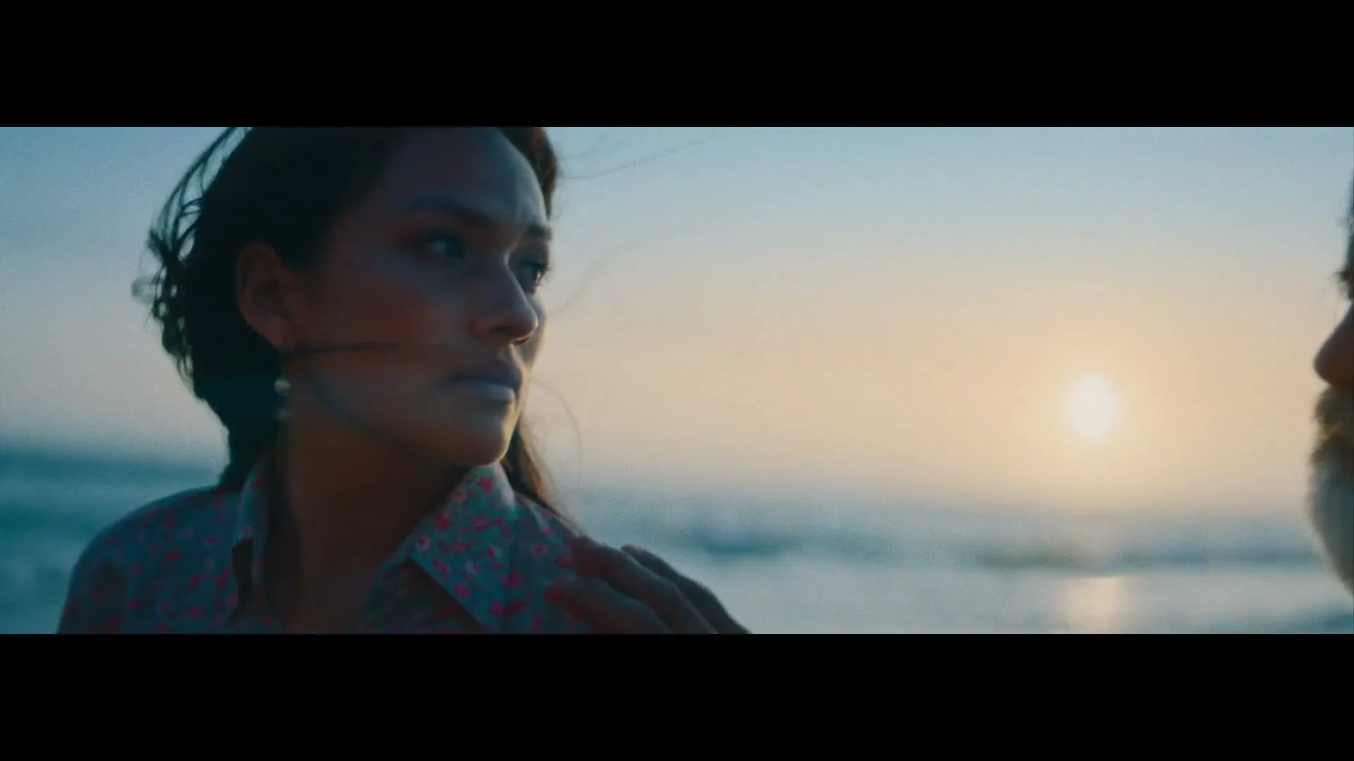 A man and a woman stand facing each other on a beach at sunset, with the ocean and sun in the background.