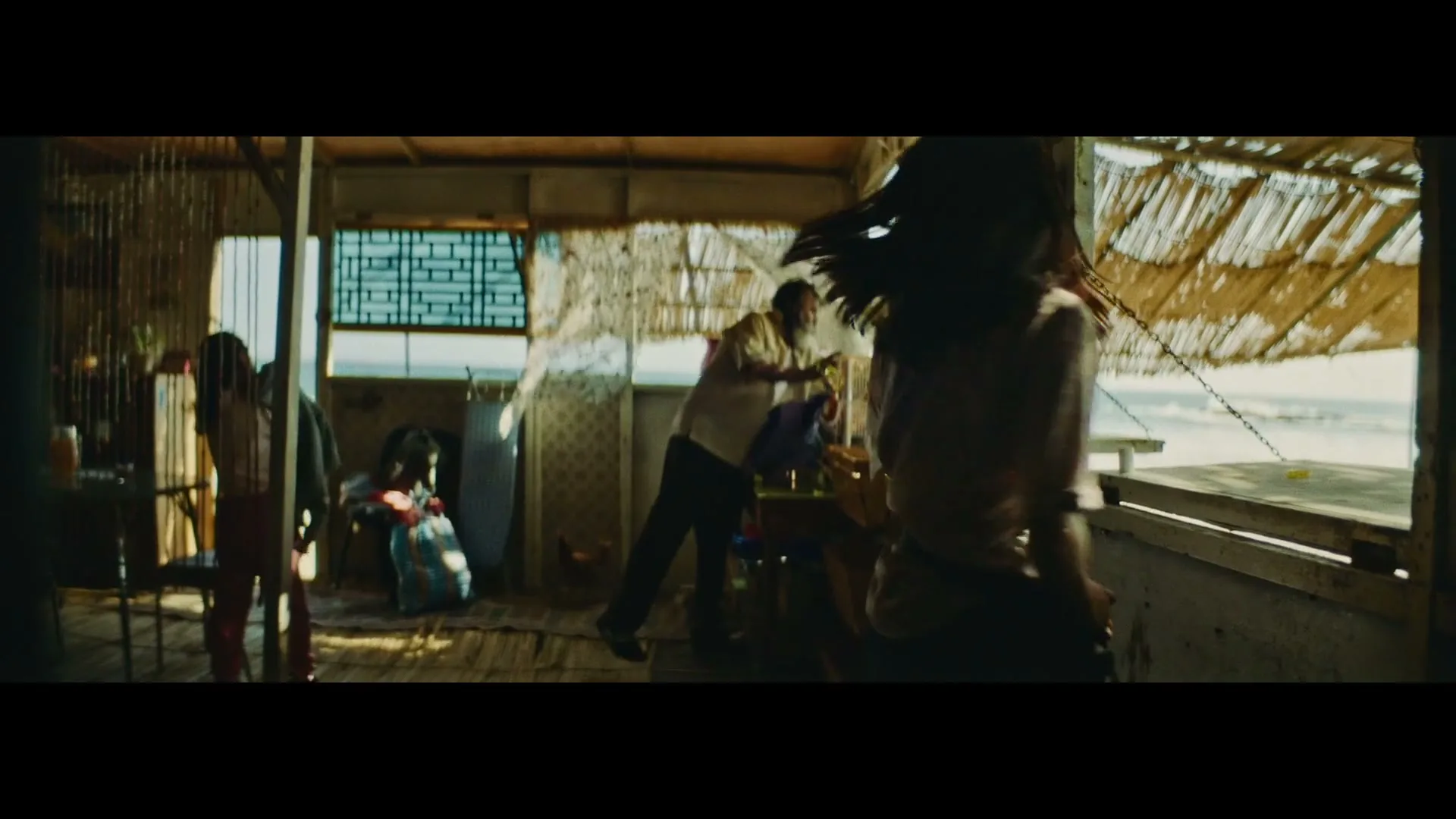 A young woman in a patterned shirt and jeans walks towards an older man in a white tank top and dark pants, who is standing in a rustic, sunlit shack by the ocean.
