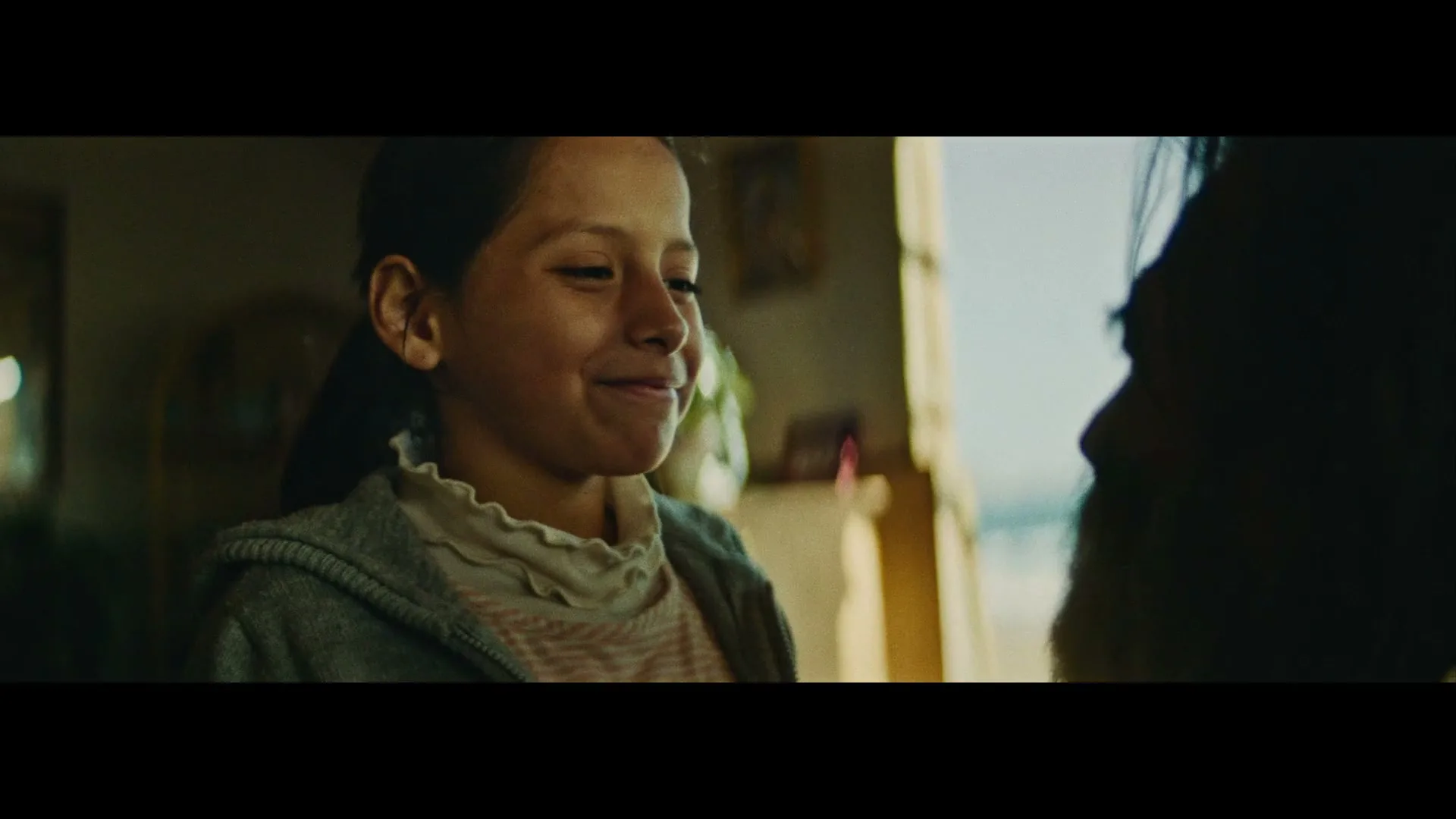 A young girl with dark hair smiles warmly, looking at an adult whose silhouette is visible on the right, in a dimly lit interior.