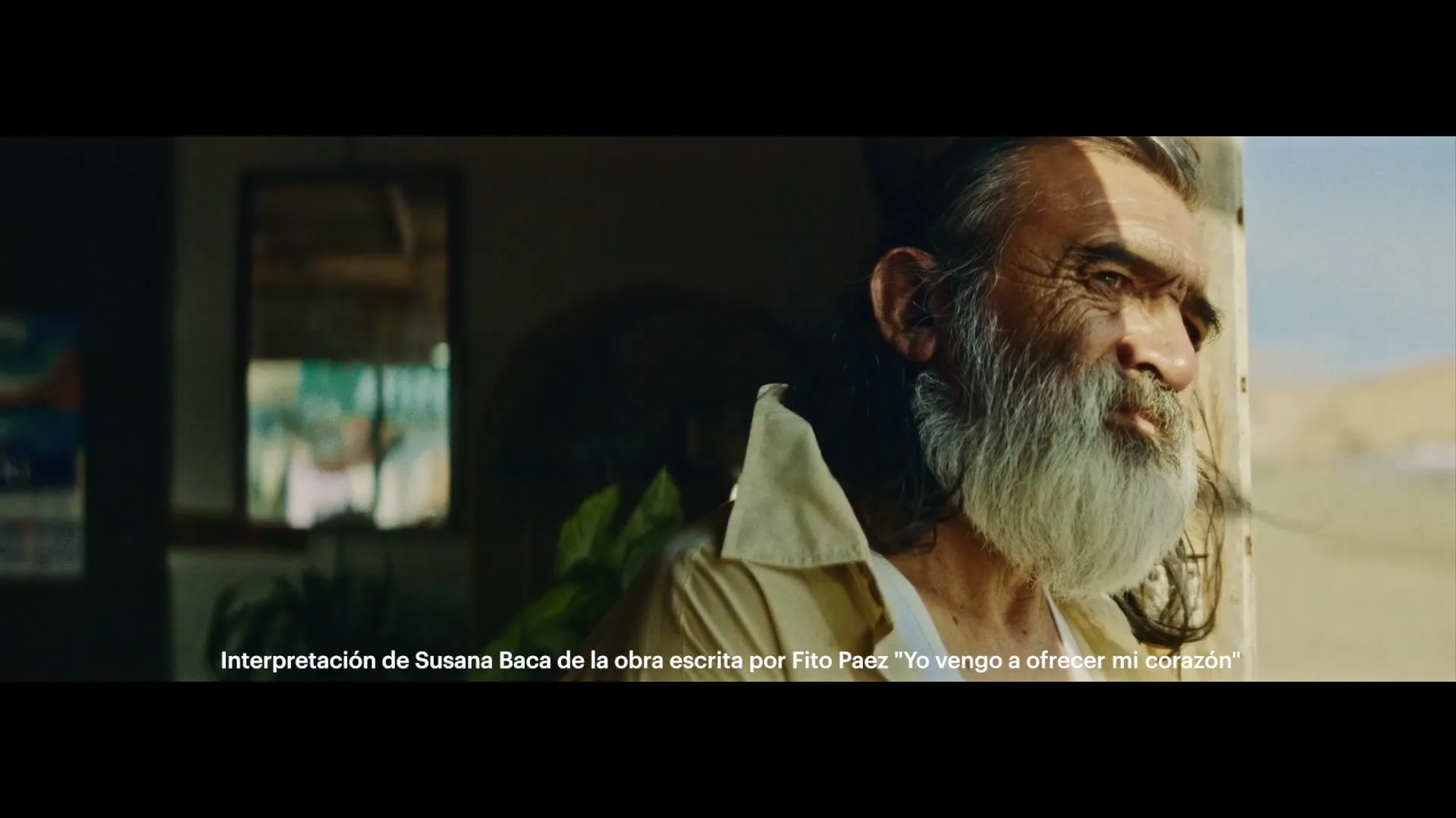 A man with a long white beard and dark hair looks out a window, with sunlight on his face and a dark room behind him.