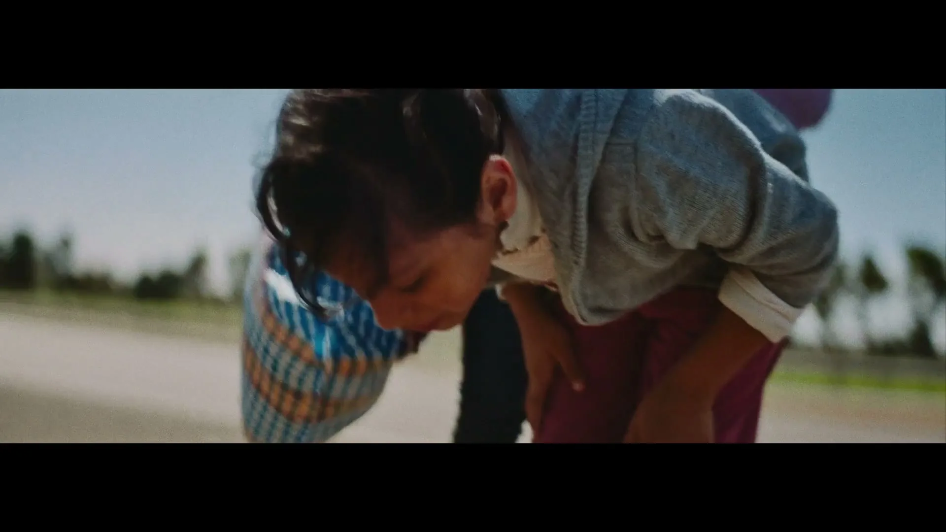 A young girl with dark hair in a grey hoodie bends down to pick something up from the ground, with another person behind her, outdoors on a sunny day.